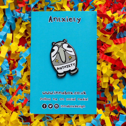 Antxiety Enamel Pin - Anxiety on blue backing card on a blue, yellow and red card confetti background. The pin is a grey and white anteater with yellow lightning bolts and the word antxiety across its belly. The pin is designed to raise awareness for anxiety disorders and anxious emotions.