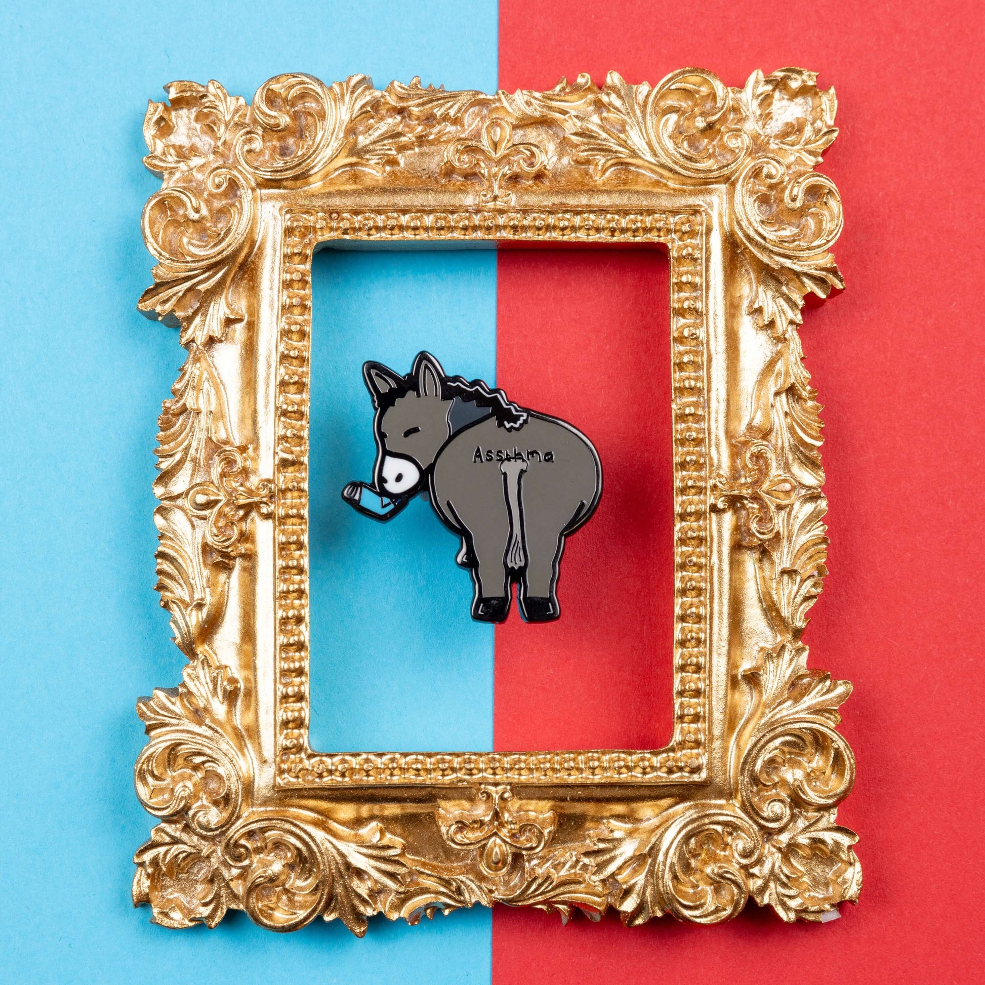 Assthma Enamel Pin - Asthma on a blue and red card background inside a gold ornate frame. A grey donkey ass showing its behind with a blue asthma pump in its mouth and the word Assthma across its behind. The pin is designed to raise awareness for asthma.