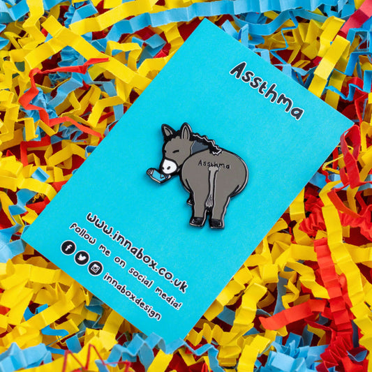 Assthma Enamel Pin - Asthma on blue backing card laid on a blue, yellow and red card confetti background. A grey donkey ass showing its behind with a blue asthma pump in its mouth and the word Assthma across its behind. The pin is designed to raise awareness for asthma.