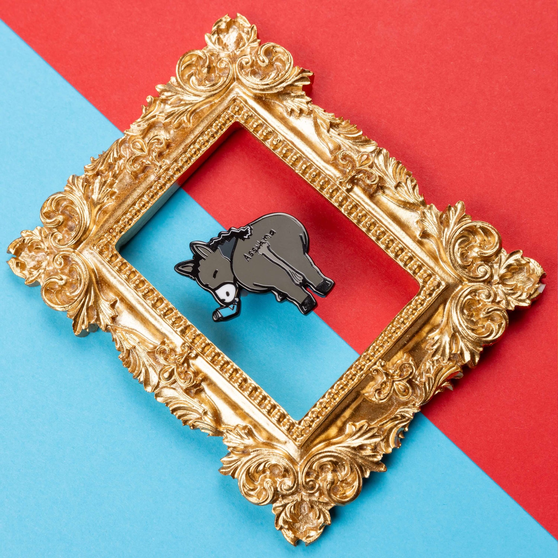 Assthma Enamel Pin - Asthma on a blue and red card background inside a gold ornate frame. A grey donkey ass showing its behind with a blue asthma pump in its mouth and the word Assthma across its behind. The pin is designed to raise awareness for asthma.