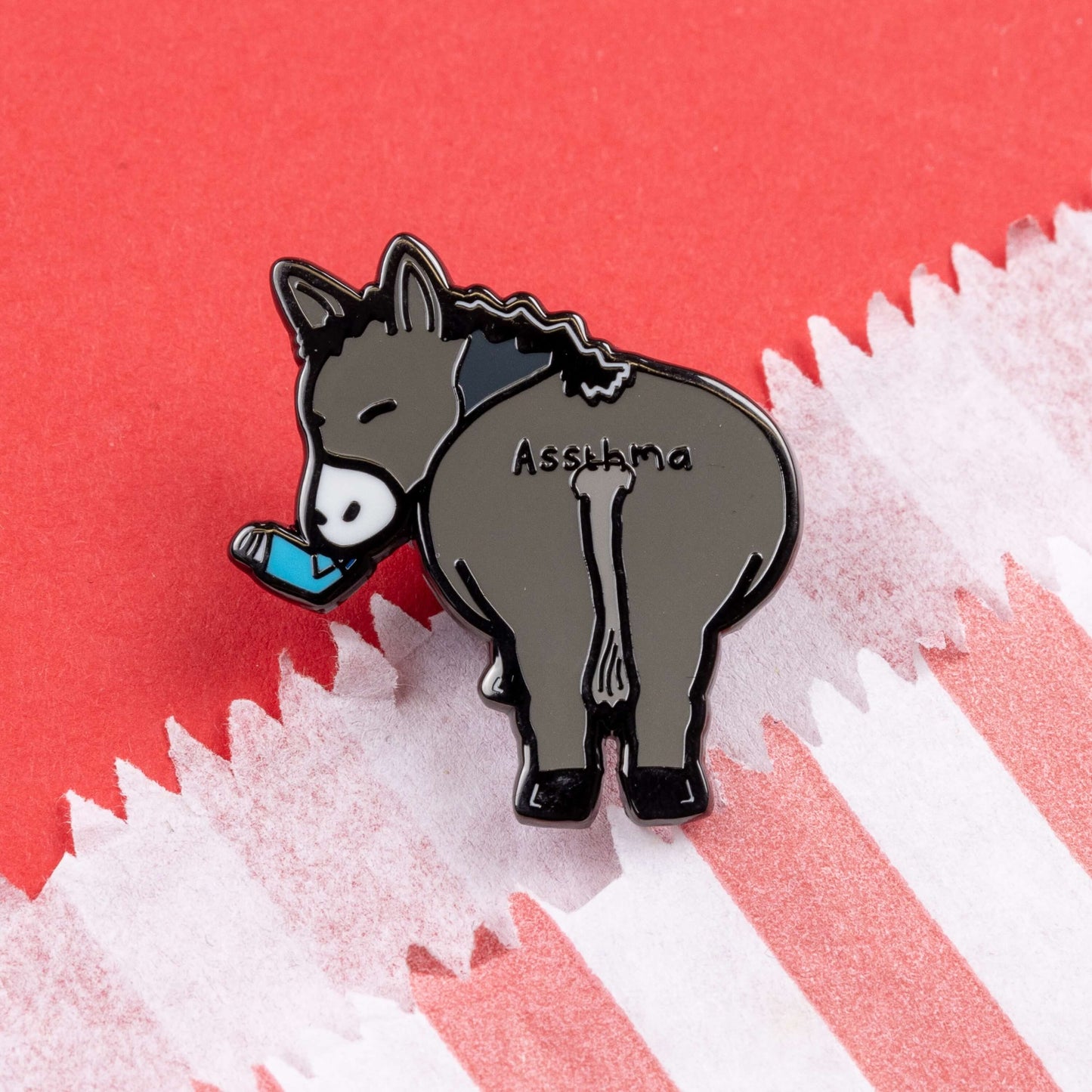 Assthma Enamel Pin - Asthma on a red card background. A grey donkey ass showing its behind with a blue asthma pump in its mouth and the word Assthma across its behind. The pin is designed to raise awareness for asthma.