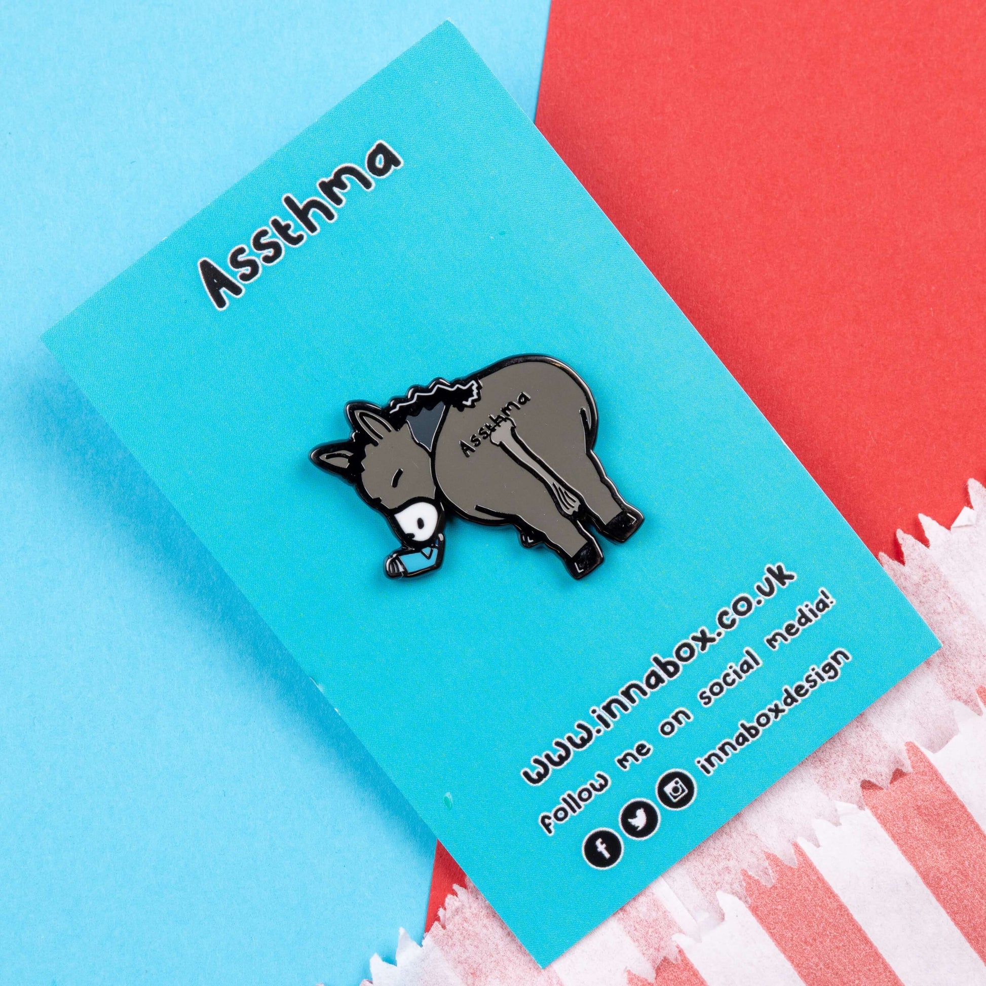 Assthma Enamel Pin - Asthma on blue backing card laid on a blue and red card background. A grey donkey ass showing its behind with a blue asthma pump in its mouth and the word Assthma across its behind. The pin is designed to raise awareness for asthma.