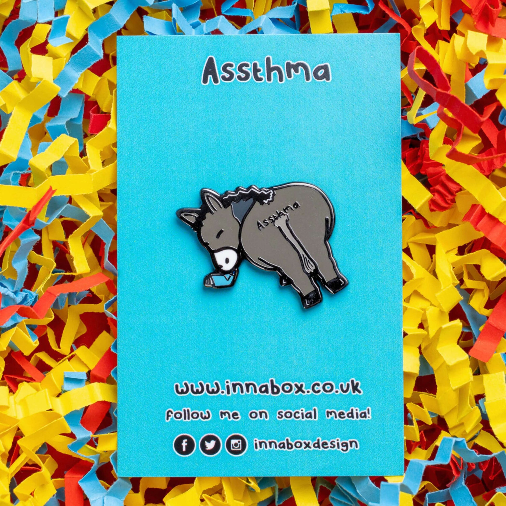 Assthma Enamel Pin - Asthma on blue backing card laid on a blue, yellow and red card confetti background. A grey donkey ass showing its behind with a blue asthma pump in its mouth and the word Assthma across its behind. The pin is designed to raise awareness for asthma.