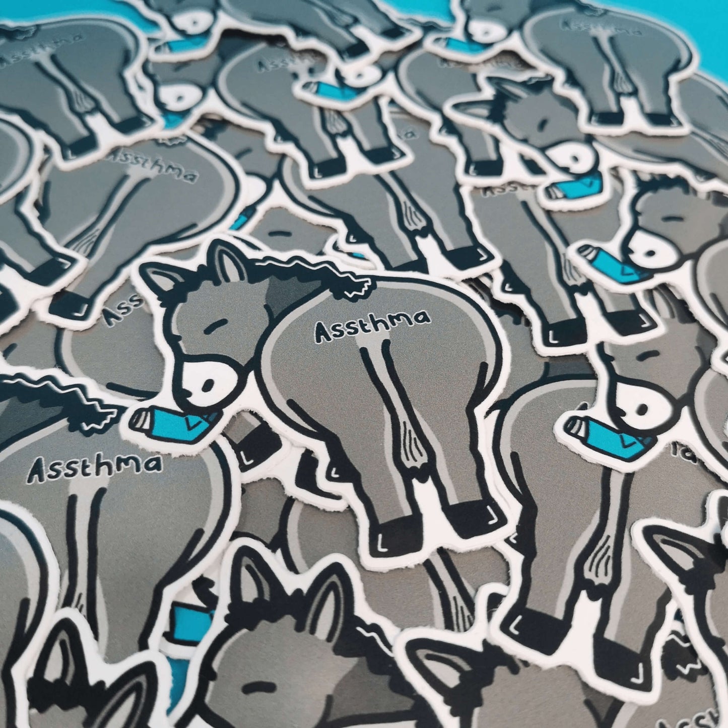 Assthma Sticker - Asthma shown laying over multiple stickers. A grey donkey ass showing its behind with a blue asthma pump in its mouth and the word Assthma across its behind. The sticker is designed to raise awareness for asthma.