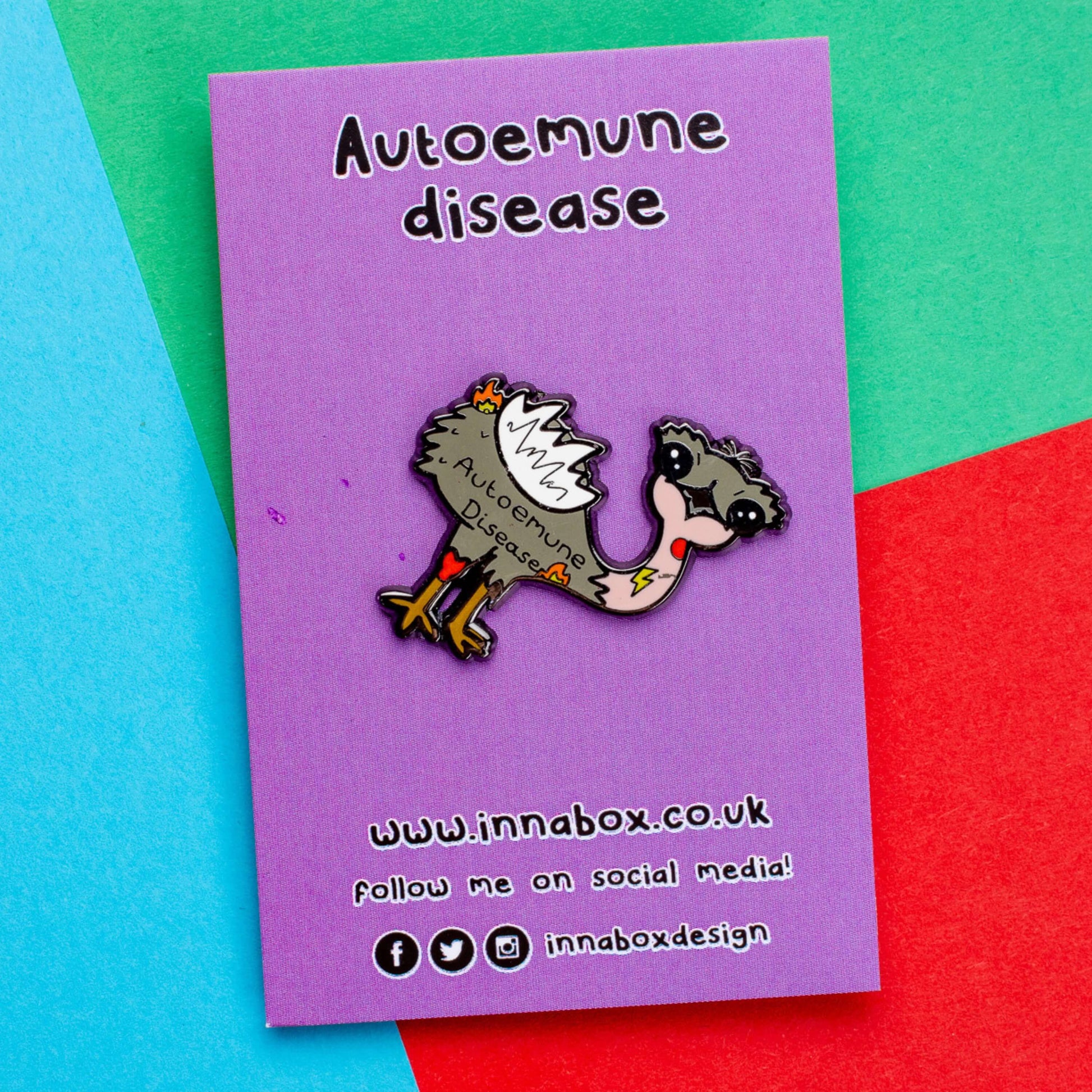 Autoemune Enamel Pin - Autoimmune Disease on a purple backing card laid on a red, blue and green background. The pin is a grey and white emu bird with various problems highlighted with lightning bolts, flames and red circles with the words autoemune disease written across its belly. Design created to raise awareness for autoimmune diseases.