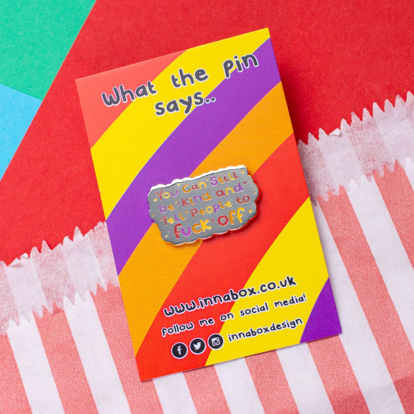The You Can Still be Kind and Tell People to F**k Off Enamel Pin on red, yellow, purple and orange stripe backing card with top text reading 'what the pin says..' laid on a red, blue and green card background. The shiny silver enamel pin has rainbow writing reading 'you can still be kind and tell people to fuck off' with multicoloured sparkles, dots and stars.