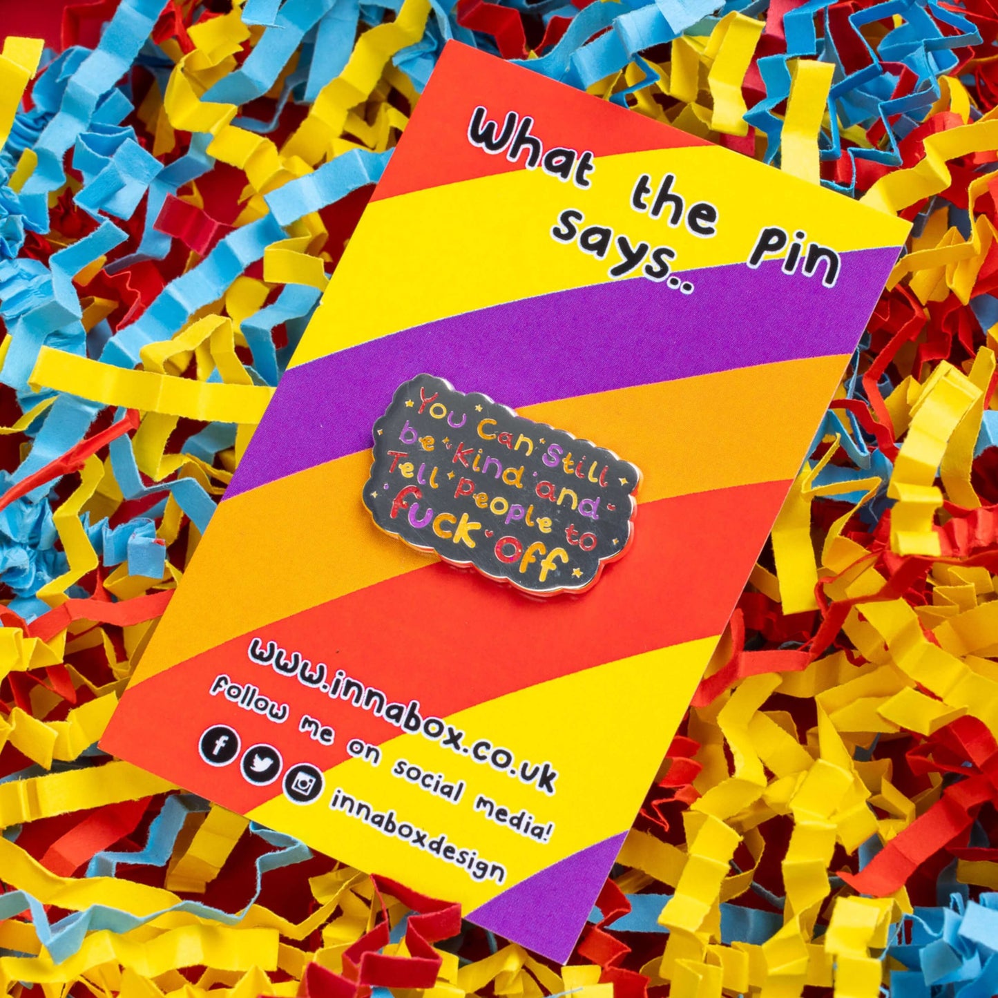 The You Can Still be Kind and Tell People to F**k Off Enamel Pin on red, yellow, purple and orange stripe backing card with top text reading 'what the pin says..' laid on a red, blue and yellow card confetti background. The shiny silver enamel pin has rainbow writing reading 'you can still be kind and tell people to fuck off' with multicoloured sparkles, dots and stars.