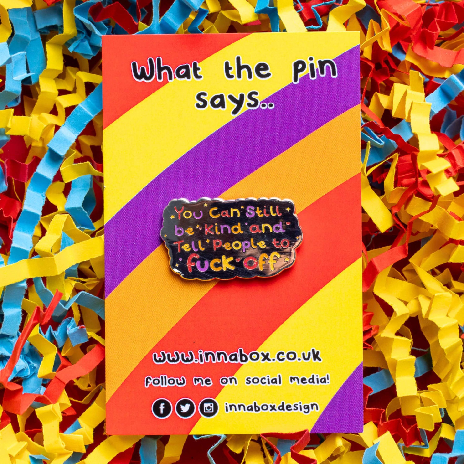 The You Can Still be Kind and Tell People to F**k Off Enamel Pin on red, yellow, purple and orange stripe backing card with top text reading 'what the pin says..' laid on a red, blue and yellow card confetti background. The shiny silver enamel pin has rainbow writing reading 'you can still be kind and tell people to fuck off' with multicoloured sparkles, dots and stars.