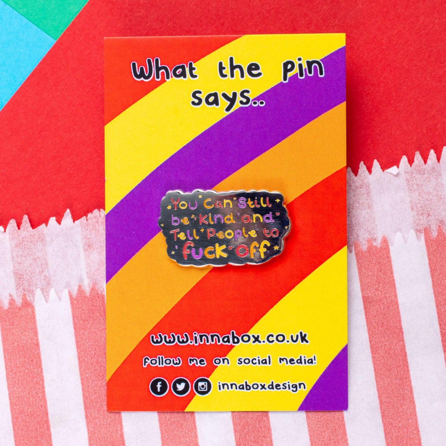 The You Can Still be Kind and Tell People to F**k Off Enamel Pin on red, yellow, purple and orange stripe backing card with top text reading 'what the pin says..' laid on a red, blue and green card background. The shiny silver enamel pin has rainbow writing reading 'you can still be kind and tell people to fuck off' with multicoloured sparkles, dots and stars.