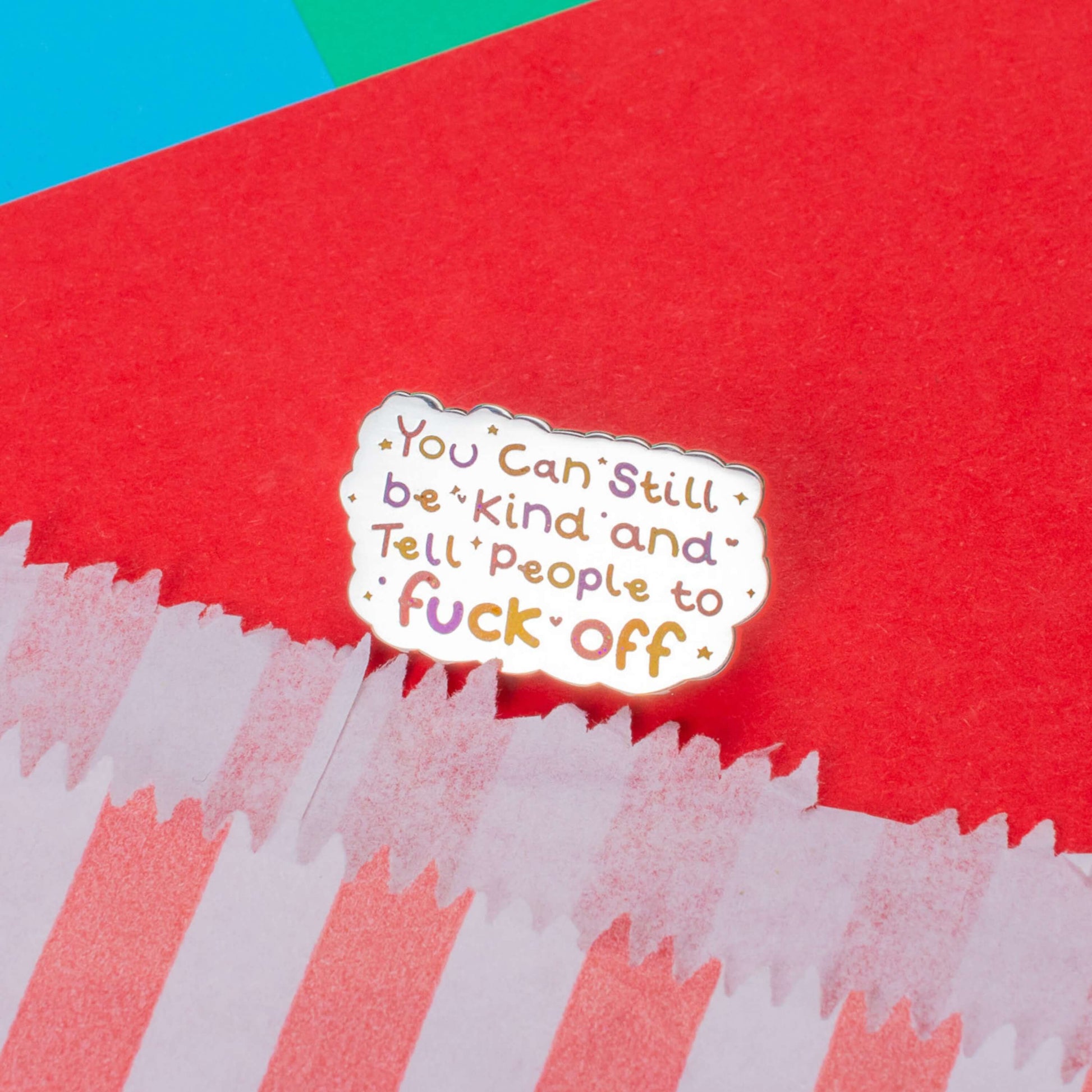 The You Can Still be Kind and Tell People to F**k Off Enamel Pin on a red, blue and green card background. The shiny silver enamel pin has rainbow writing reading 'you can still be kind and tell people to fuck off' with multicoloured sparkles, dots and stars.