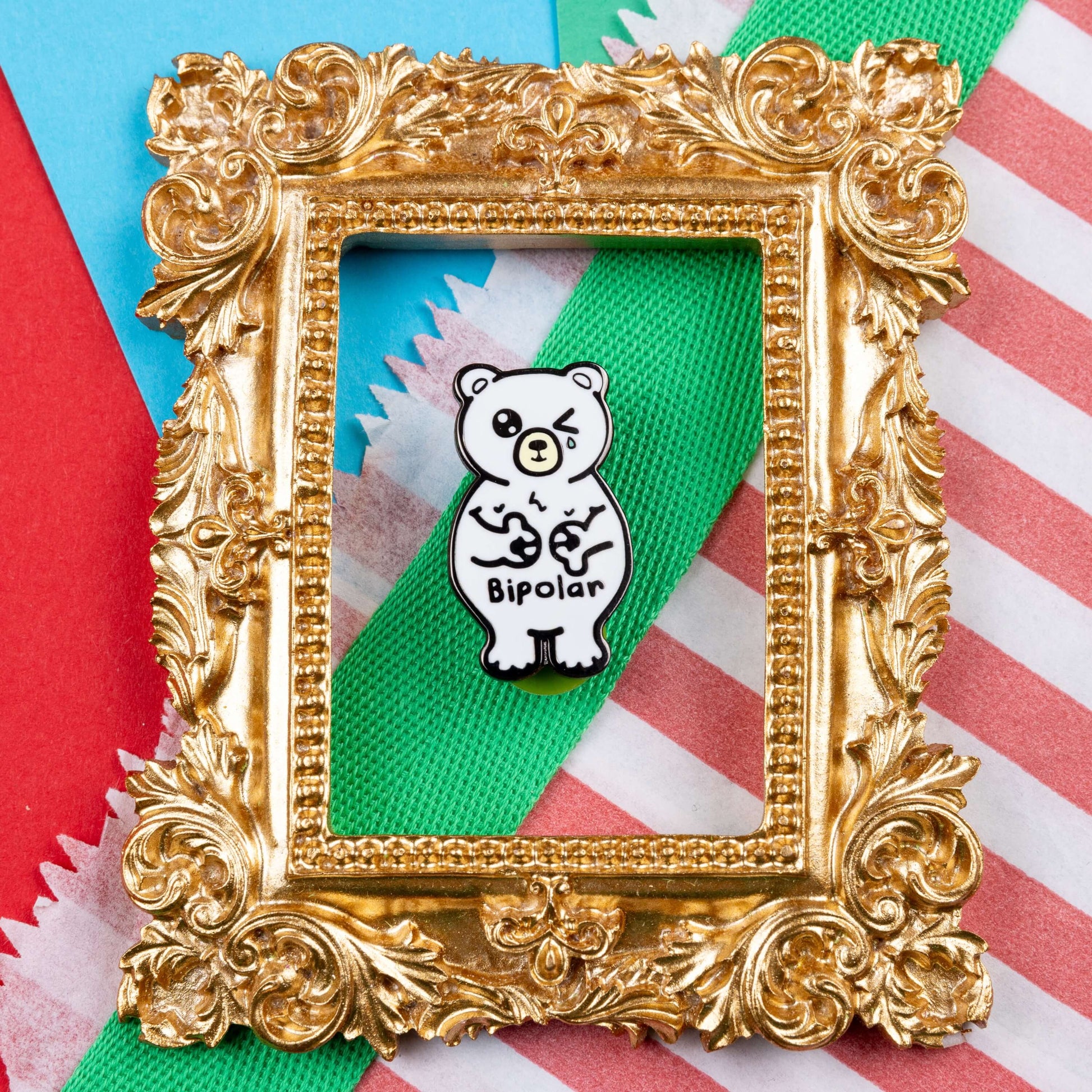 Bipolar Bear Enamel Pin - Bipolar on a blue, red and green card background in a gold ornate frame. The pin is a white polar bear with one eye closed crying whilst smiling clutching its chest, across its tummy reads bipolar. The pun pin is raising awareness for bipolar disorder.