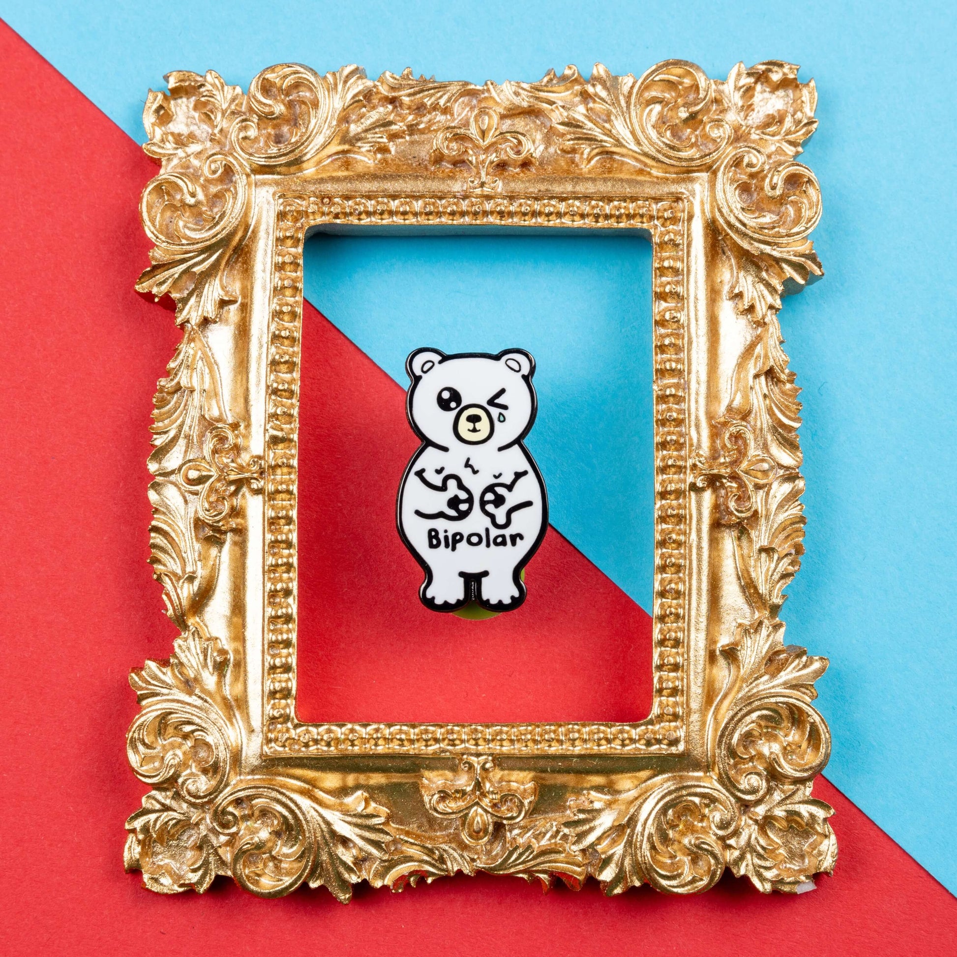 Bipolar Bear Enamel Pin - Bipolar on a blue and red card background in a gold ornate frame. The pin is a white polar bear with one eye closed crying whilst smiling clutching its chest, across its tummy reads bipolar. The pun pin is raising awareness for bipolar disorder.