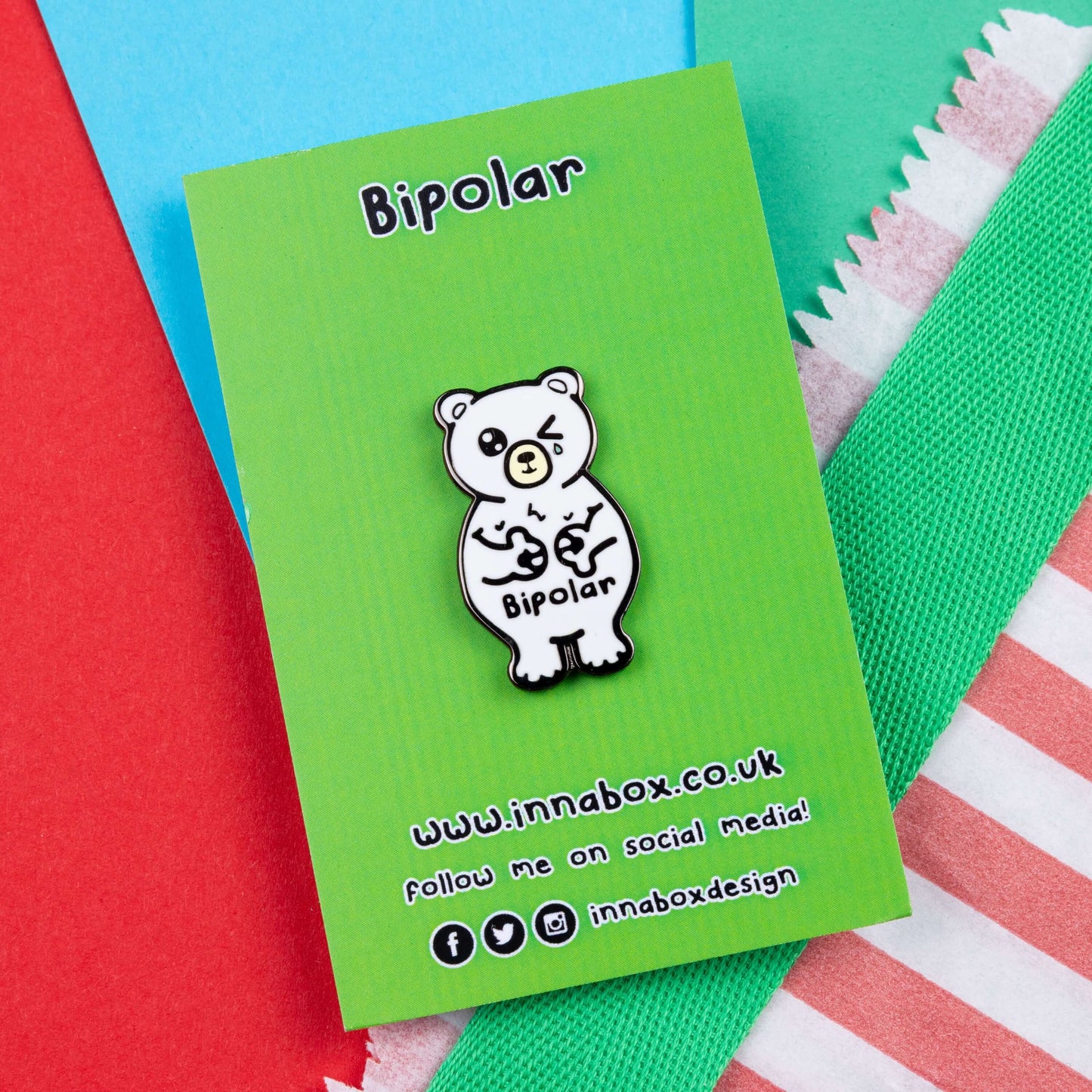 Bipolar Bear Enamel Pin - Bipolar on its green backing card laid on a blue, green and red card background. The pin is a white polar bear with one eye closed crying whilst smiling clutching its chest, across its tummy reads bipolar. The pun pin is raising awareness for bipolar disorder.