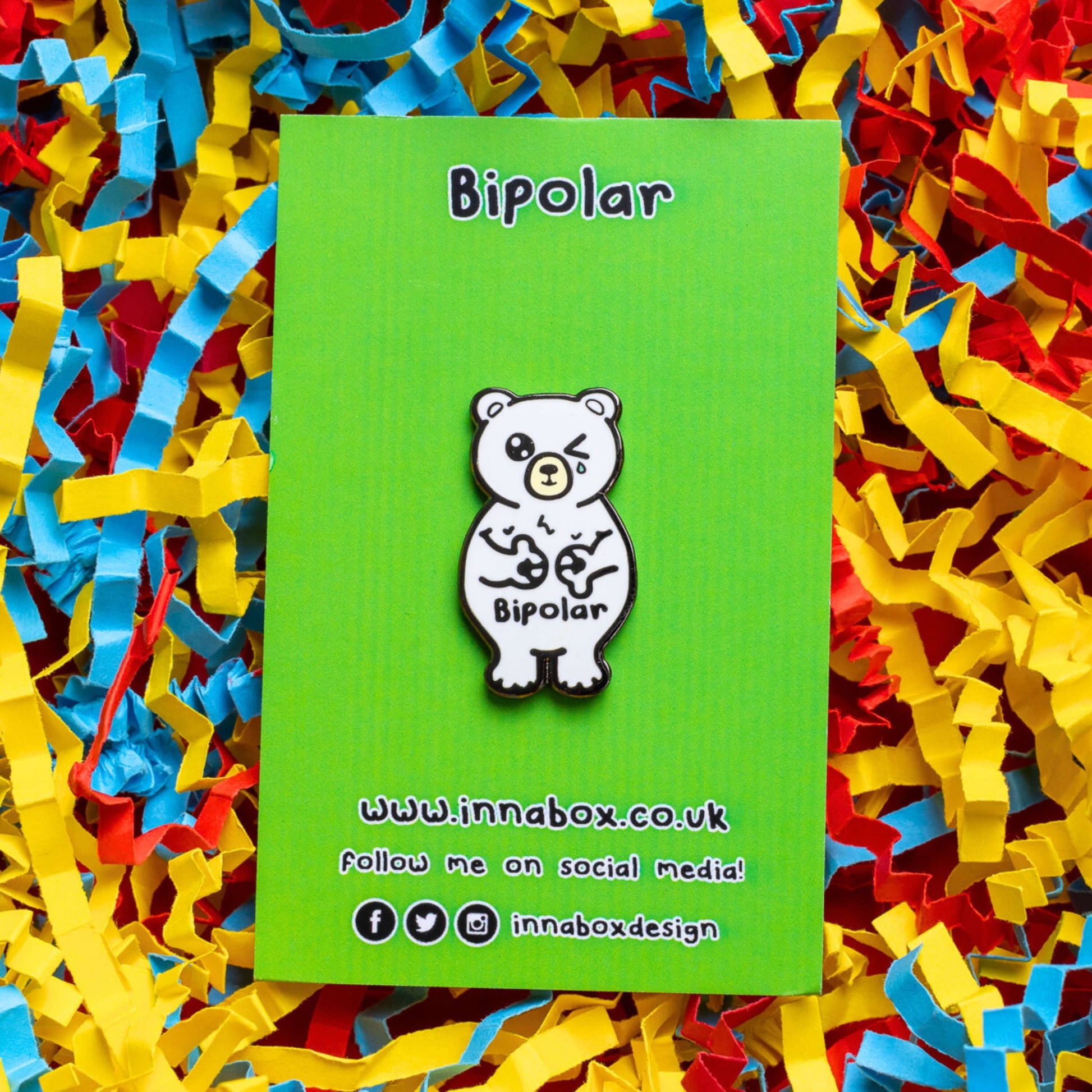 Bipolar Bear Enamel Pin - Bipolar on its green backing card laid on a blue, yellow and red card confetti background. The pin is a white polar bear with one eye closed crying whilst smiling clutching its chest, across its tummy reads bipolar. The pun pin is raising awareness for bipolar disorder.