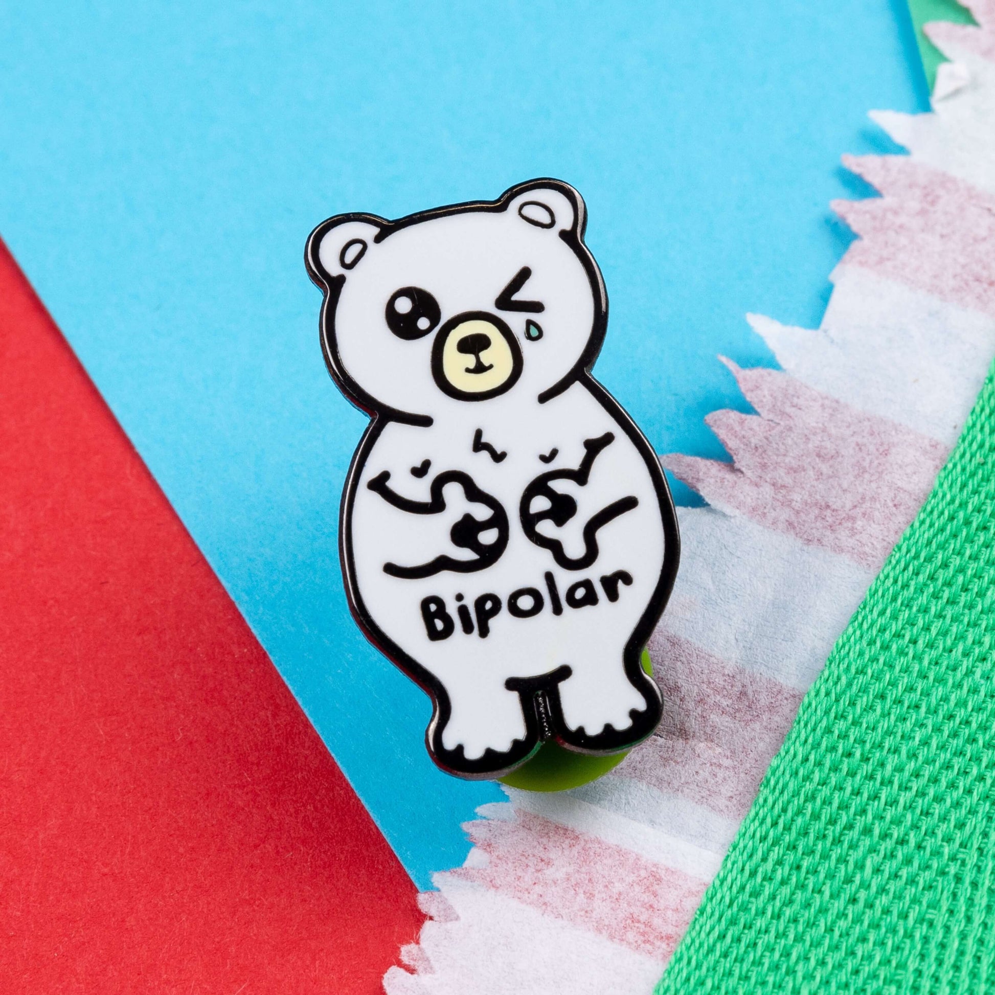 Bipolar Bear Enamel Pin - Bipolar on a blue and red card background. The pin is a white polar bear with one eye closed crying whilst smiling clutching its chest, across its tummy reads bipolar. The pun pin is raising awareness for bipolar disorder.