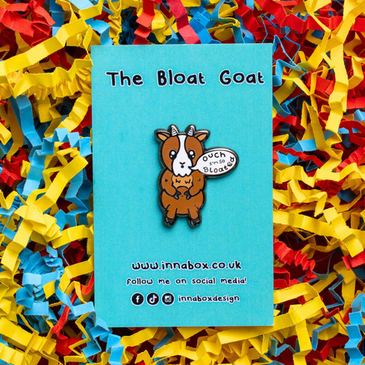 The Bloat Goat Enamel Pin on blue backing card laid on a red, yellow and blue card confetti background. The brown frustrated goat pin badge is stood up clutching its tummy with a speech bubble reading 'ouch I'm so bloated'. The hand drawn design is raising awareness for chronic illnesses or hidden disabilities that cause bloating.