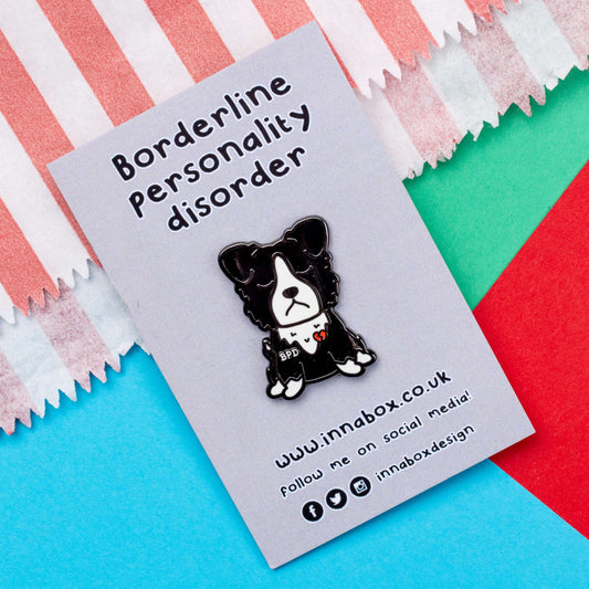 BPD Enamel Pin - Borderline Personality Disorder on grey backing card with the pin name above and the innabox social media details underneath laid on a red, blue and green card background. The black and white sad fluffy dog pin has a broken heart with BPD written across its chest. The pin is designed to raise awareness for Borderline Personality Disorder.