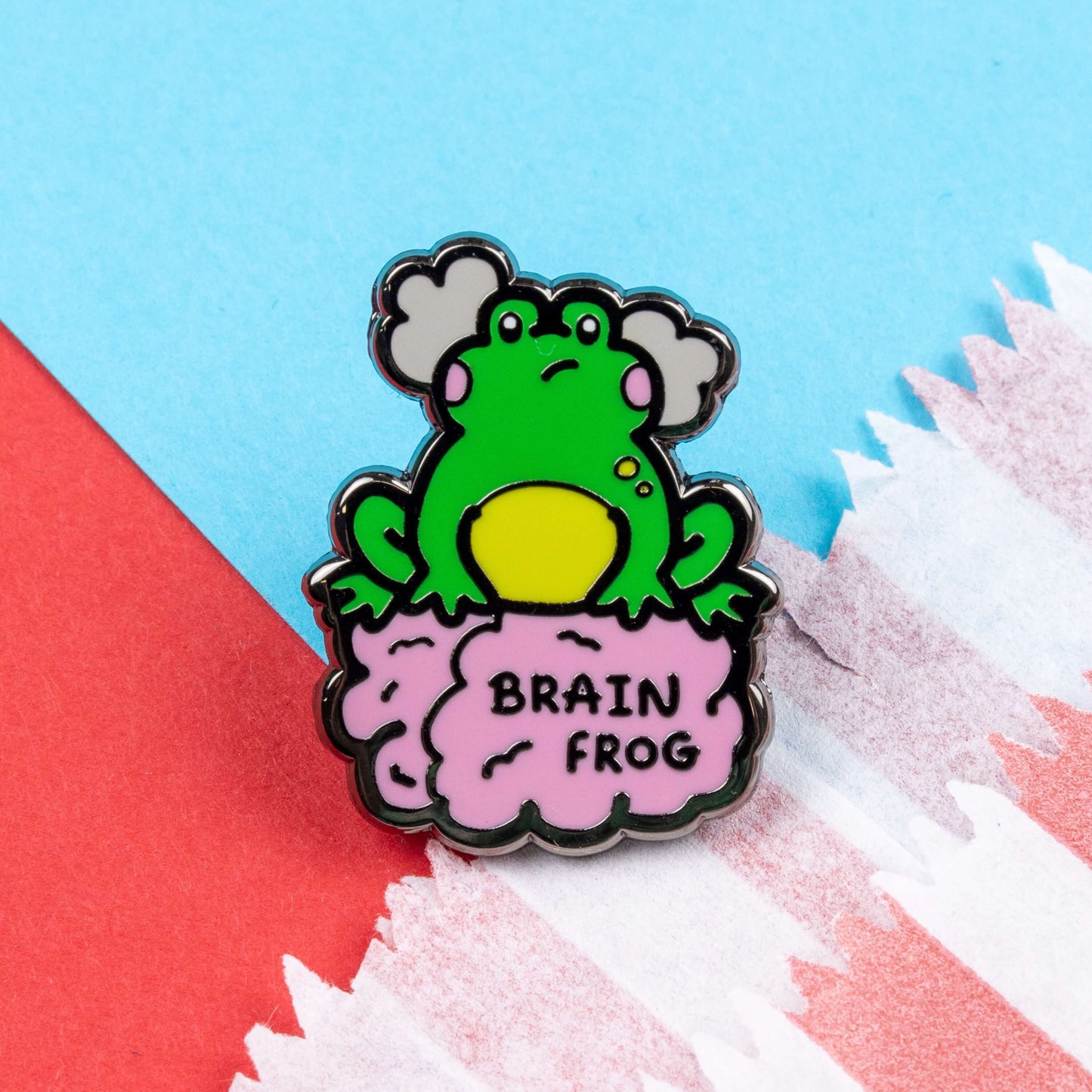 The Brain Frog Enamel Pin - Brain Fog on a blue and red card background. The pin is of a green frog with pink blush cheeks looking confused with two grey clouds by its head, it is sat on a pink brain with the text reading Brain Frog. The design is raising awareness for brain fog.