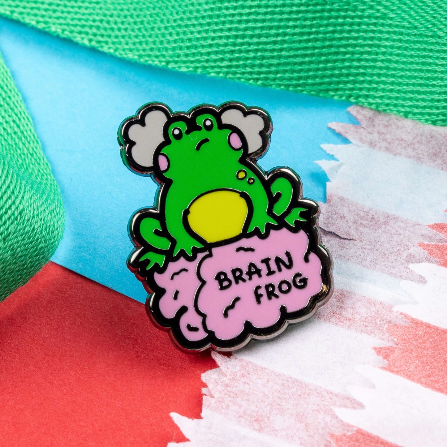 The Brain Frog Enamel Pin - Brain Fog on a blue and red card background with a green ribbon. The pin is of a green frog with pink blush cheeks looking confused with two grey clouds by its head, it is sat on a pink brain with the text reading Brain Frog. The design is raising awareness for brain fog.