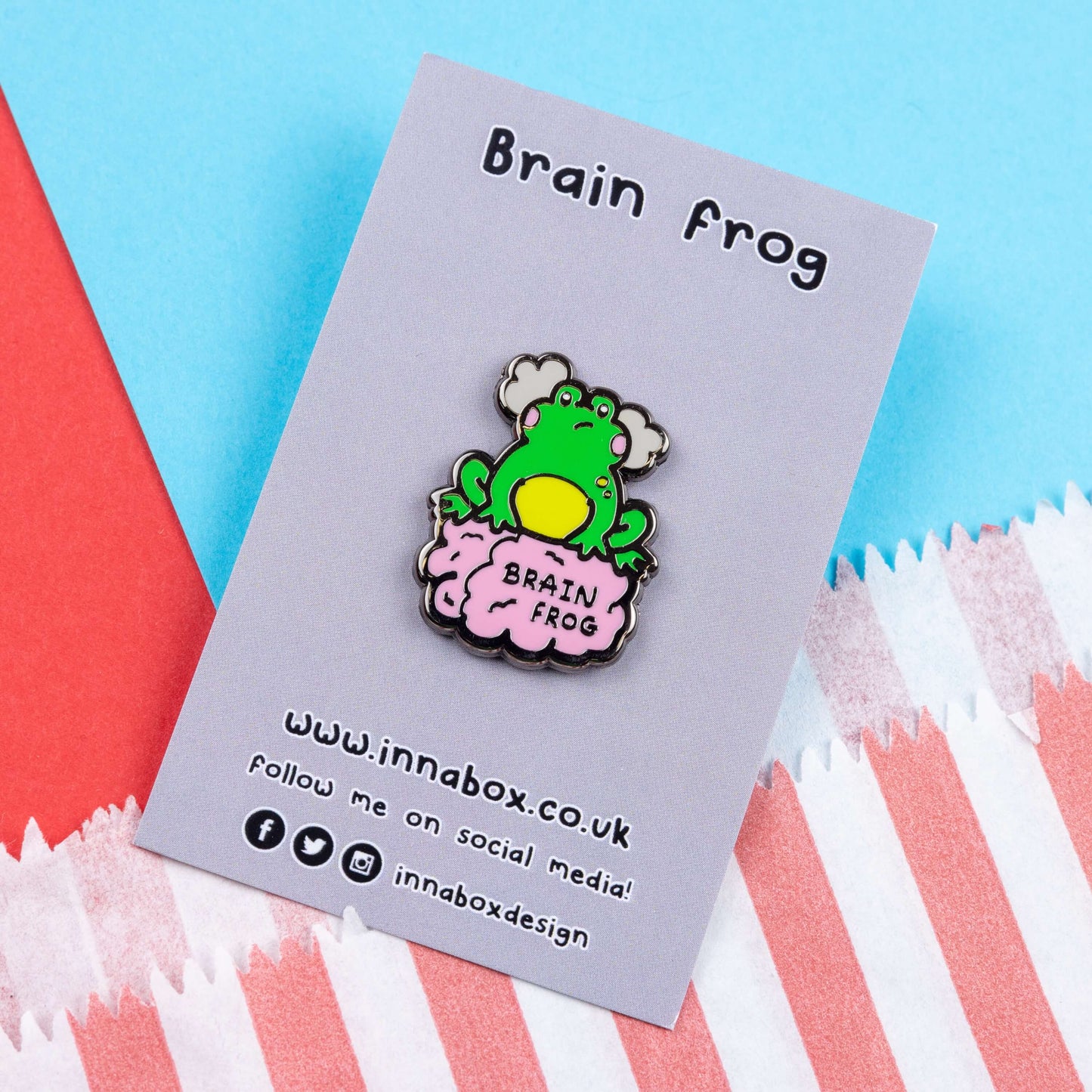 The Brain Frog Enamel Pin - Brain Fog on its grey backing card laid on a blue and red card background. The pin is of a green frog with pink blush cheeks looking confused with two grey clouds by its head, it is sat on a pink brain with the text reading Brain Frog. The design is raising awareness for brain fog.