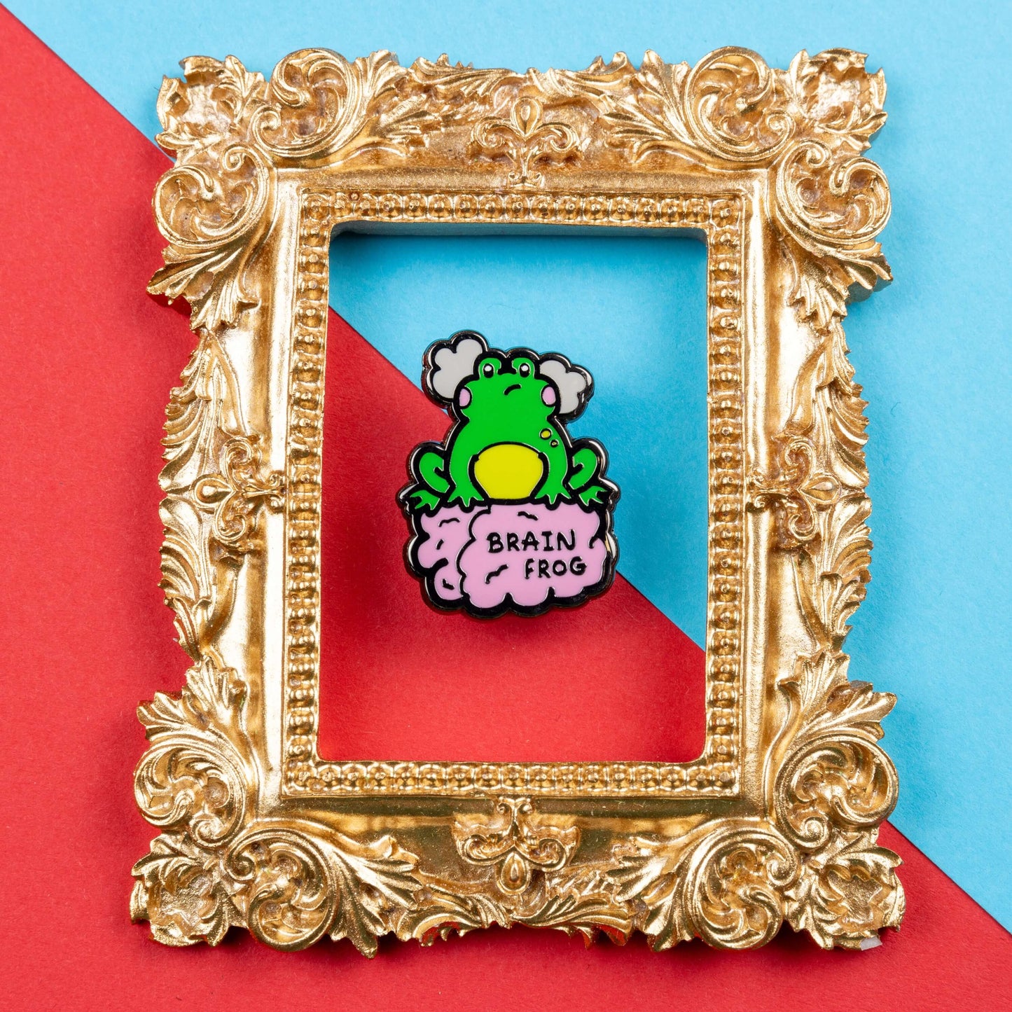 The Brain Frog Enamel Pin - Brain Fog on a blue and red card background inside a gold ornate frame. The pin is of a green frog with pink blush cheeks looking confused with two grey clouds by its head, it is sat on a pink brain with the text reading Brain Frog. The design is raising awareness for brain fog.