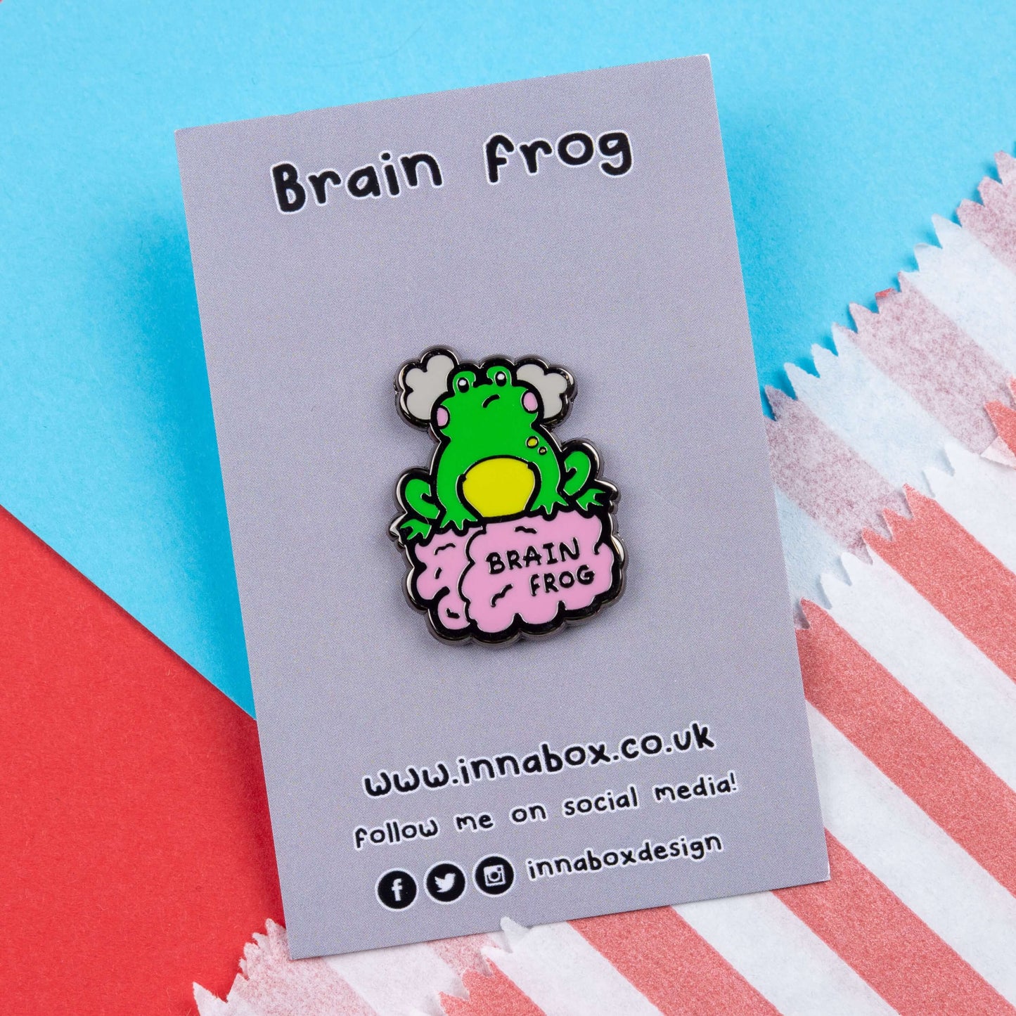 The Brain Frog Enamel Pin - Brain Fog on its grey backing card laid on a blue and red card background. The pin is of a green frog with pink blush cheeks looking confused with two grey clouds by its head, it is sat on a pink brain with the text reading Brain Frog. The design is raising awareness for brain fog.