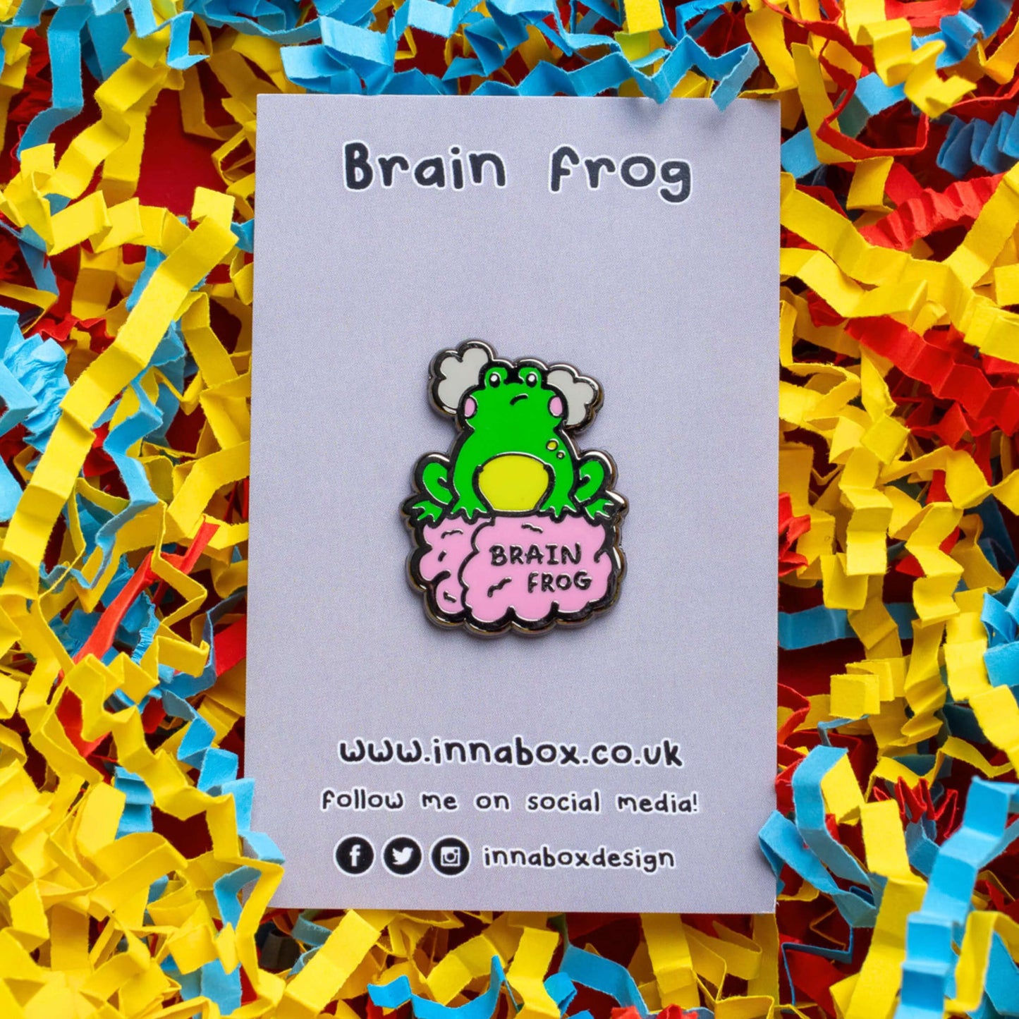 The Brain Frog Enamel Pin - Brain Fog on its grey backing card laid on a blue, yellow and red confetti card background. The pin is of a green frog with pink blush cheeks looking confused with two grey clouds by its head, it is sat on a pink brain with the text reading Brain Frog. The design is raising awareness for brain fog.