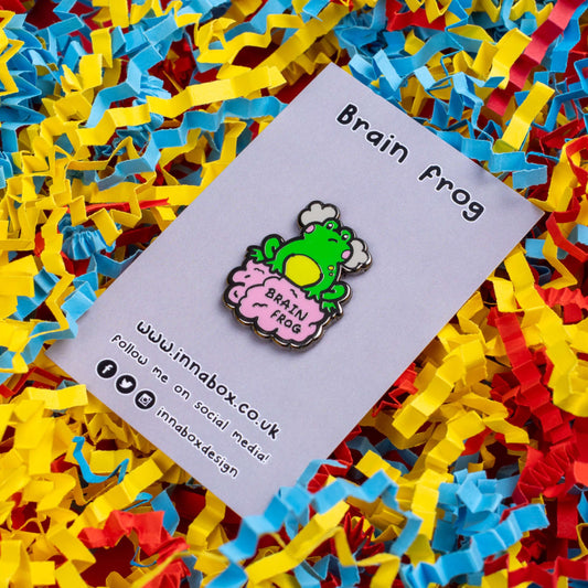 The Brain Frog Enamel Pin - Brain Fog on its grey backing card laid on a blue, yellow and red confetti card background. The pin is of a green frog with pink blush cheeks looking confused with two grey clouds by its head, it is sat on a pink brain with the text reading Brain Frog. The design is raising awareness for brain fog.