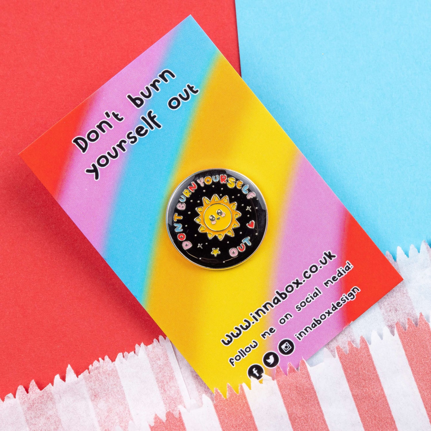 The Don't Burn Yourself Out Sunshine Enamel Pin on rainbow backing card laid on blue and red card. The circle shaped pin badge is a black base with a yellow smiling sunshine in the centre with rainbow text surrounding it reading 'don't burn yourself out' along with a red heart, yellow star and silver sparkles. The design was created as a gentle reminder to take care of yourself and self care.