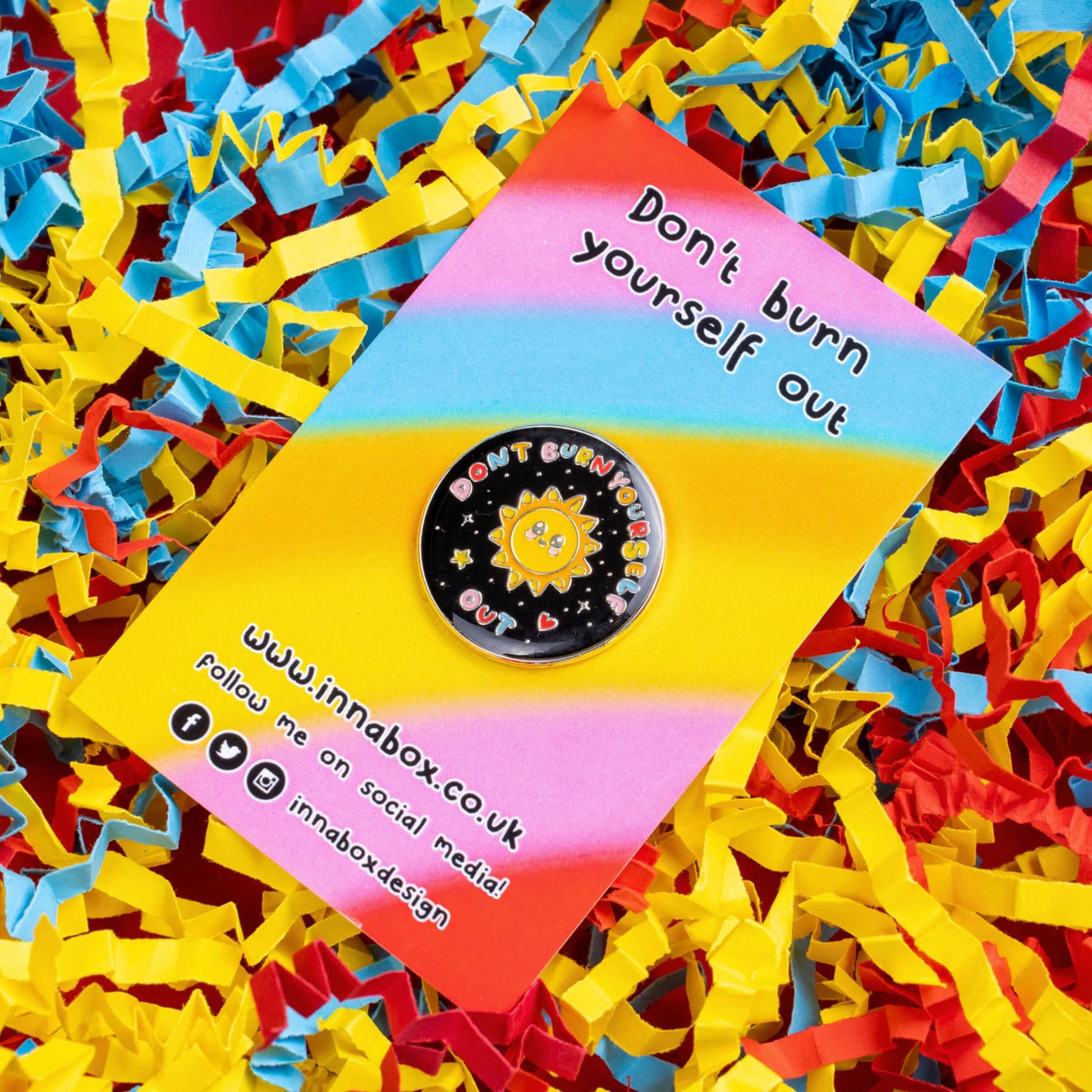 The Don't Burn Yourself Out Sunshine Enamel Pin on rainbow backing card laid on blue, yellow and red card confetti. The circle shaped pin badge is a black base with a yellow smiling sunshine in the centre with rainbow text surrounding it reading 'don't burn yourself out' along with a red heart, yellow star and silver sparkles. The design was created as a gentle reminder to take care of yourself and self care.