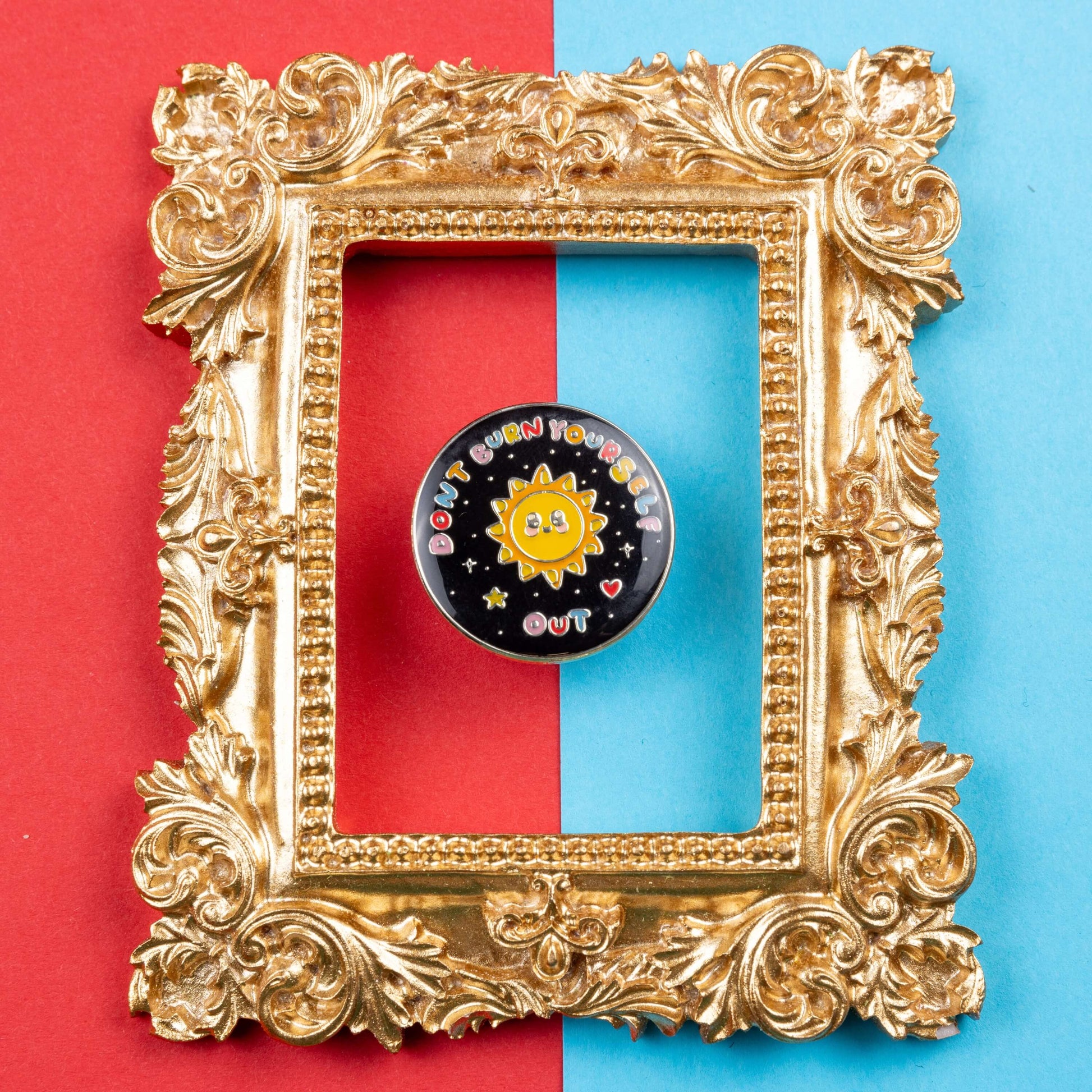 The Don't Burn Yourself Out Sunshine Enamel Pin on blue and red card in a gold ornate frame. The circle shaped pin badge is a black base with a yellow smiling sunshine in the centre with rainbow text surrounding it reading 'don't burn yourself out' along with a red heart, yellow star and silver sparkles. The design was created as a gentle reminder to take care of yourself and self care.