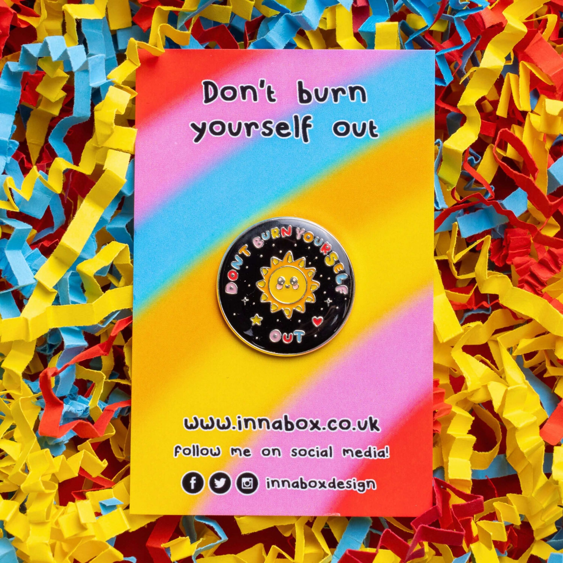 The Don't Burn Yourself Out Sunshine Enamel Pin on rainbow backing card laid on blue, yellow and red card confetti. The circle shaped pin badge is a black base with a yellow smiling sunshine in the centre with rainbow text surrounding it reading 'don't burn yourself out' along with a red heart, yellow star and silver sparkles. The design was created as a gentle reminder to take care of yourself and self care.