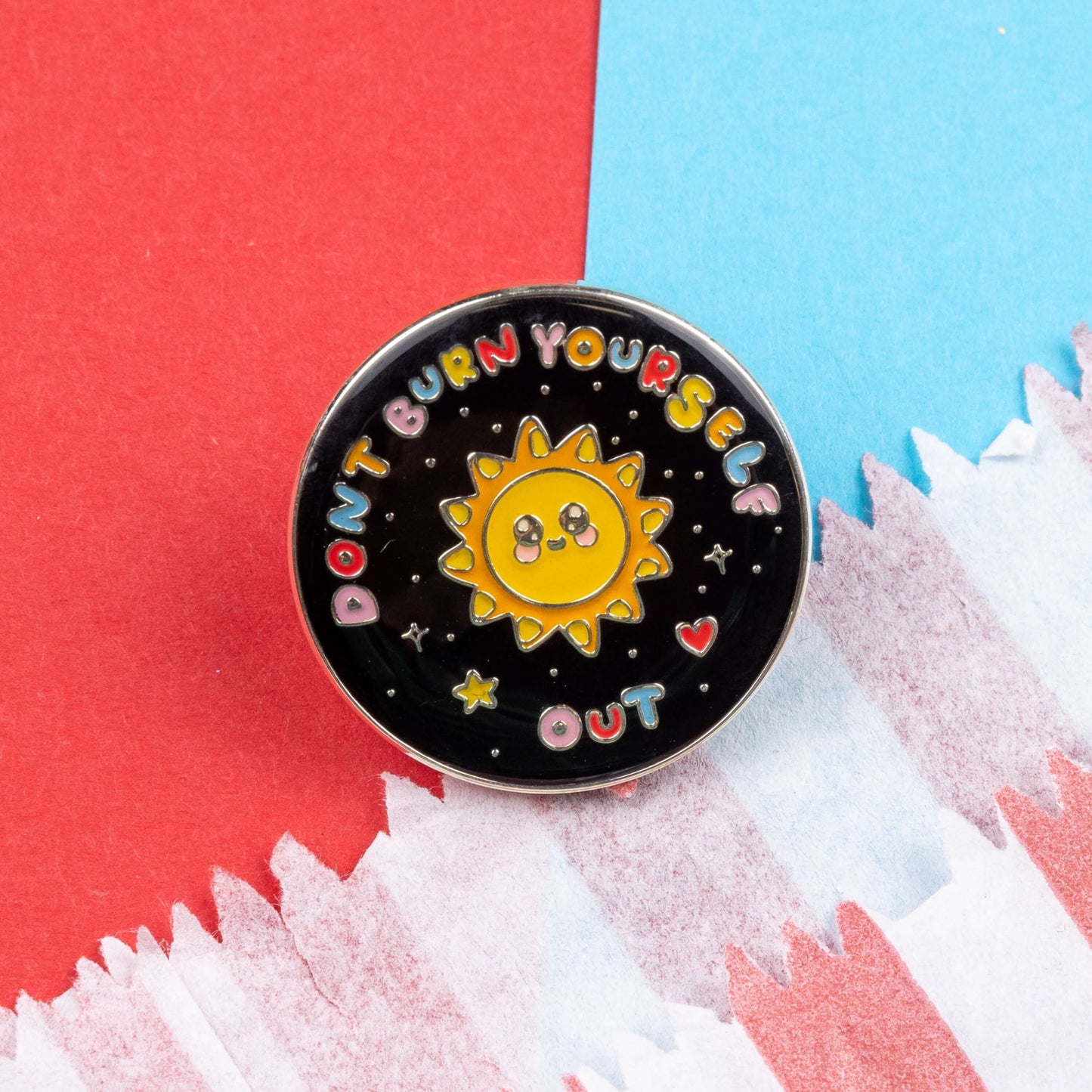 The Don't Burn Yourself Out Sunshine Enamel Pin on blue and red card. The circle shaped pin badge is a black base with a yellow smiling sunshine in the centre with rainbow text surrounding it reading 'don't burn yourself out' along with a red heart, yellow star and silver sparkles. The design was created as a gentle reminder to take care of yourself and self care.