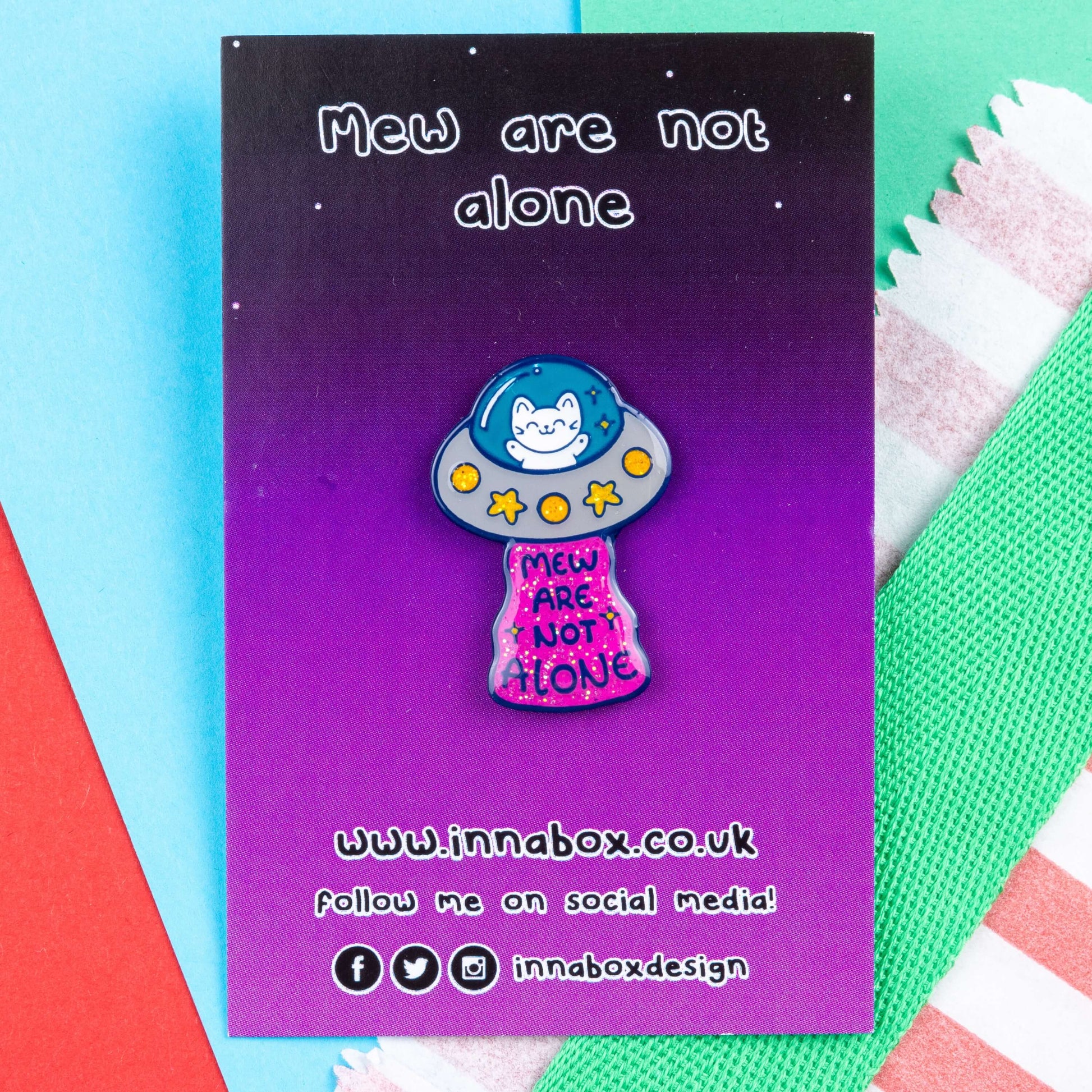 The Cat UFO Mew are not Alone Enamel Pin on its purple gradient backing card laid on a red, green and blue card background. The blue outline pin is of a white smiling cat in a silver ufo spaceship with yellow glittery stars and circles, underneath is pink glittery beam with blue text reading 'mew are not alone' a pun on you are not alone.