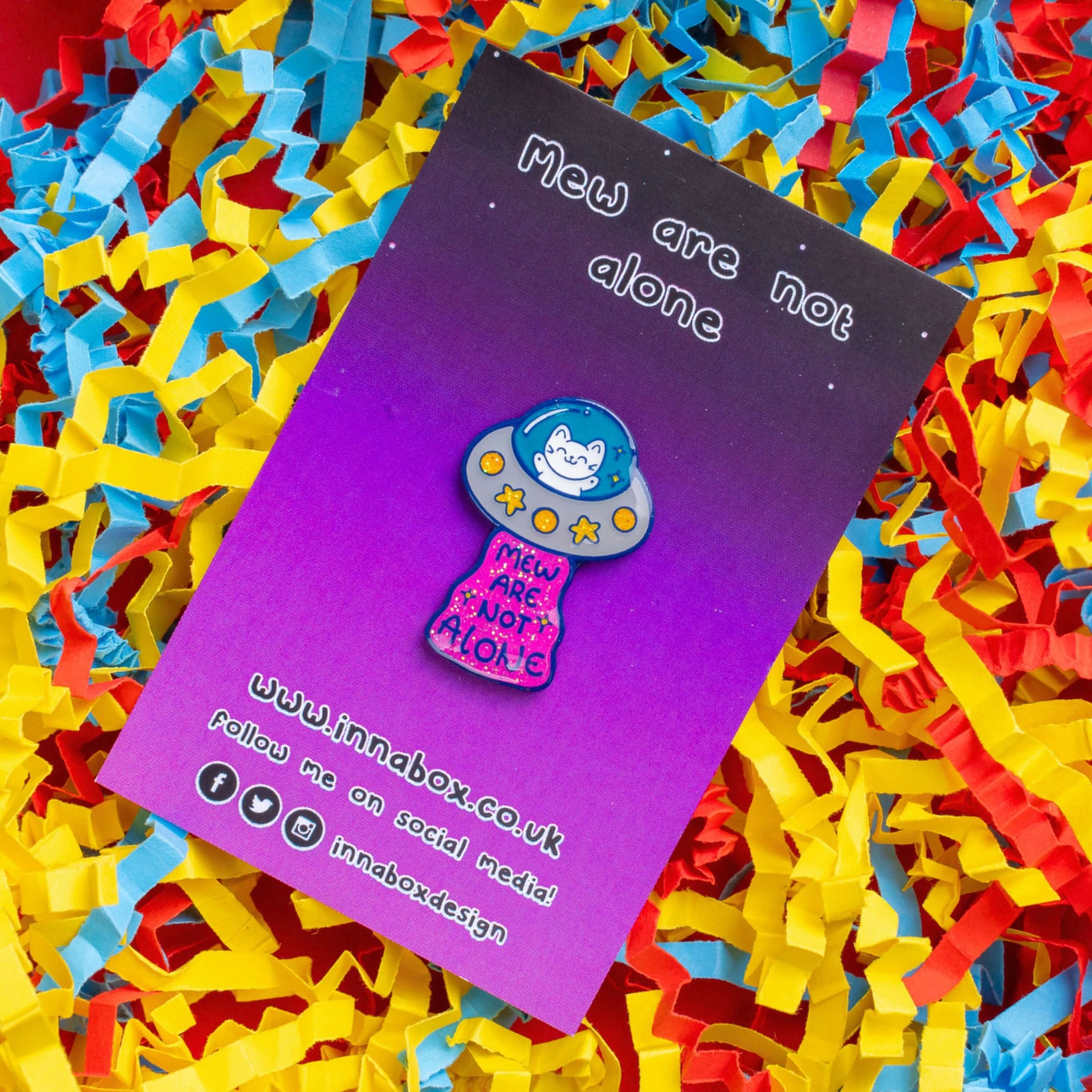 The Cat UFO Mew are not Alone Enamel Pin on its purple gradient backing card laid on a red, yellow and blue card confetti background. The blue outline pin is of a white smiling cat in a silver ufo spaceship with yellow glittery stars and circles, underneath is pink glittery beam with blue text reading 'mew are not alone' a pun on you are not alone.