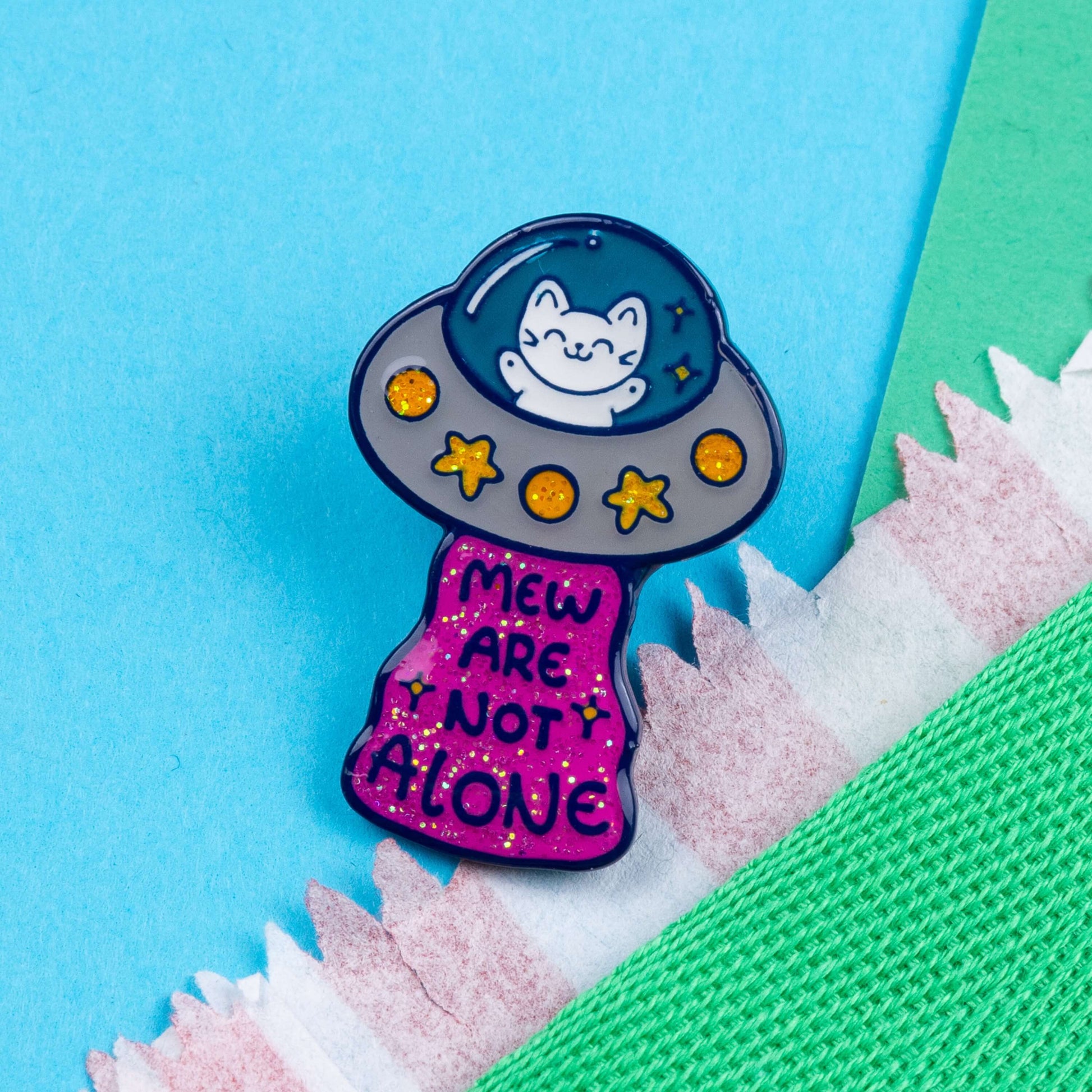 The Cat UFO Mew are not Alone Enamel Pin on a green and blue card background. The blue outline pin is of a white smiling cat in a silver ufo spaceship with yellow glittery stars and circles, underneath is pink glittery beam with blue text reading 'mew are not alone' a pun on you are not alone.