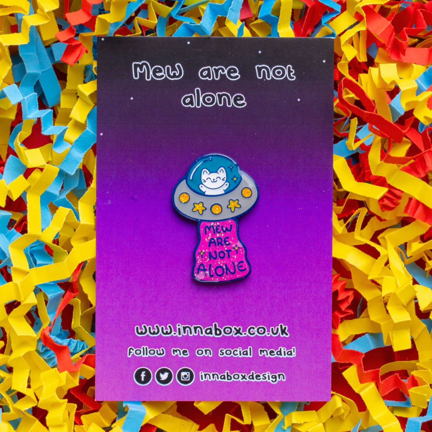 The Cat UFO Mew are not Alone Enamel Pin on its purple gradient backing card laid on a red, yellow and blue card confetti background. The blue outline pin is of a white smiling cat in a silver ufo spaceship with yellow glittery stars and circles, underneath is pink glittery beam with blue text reading 'mew are not alone' a pun on you are not alone.