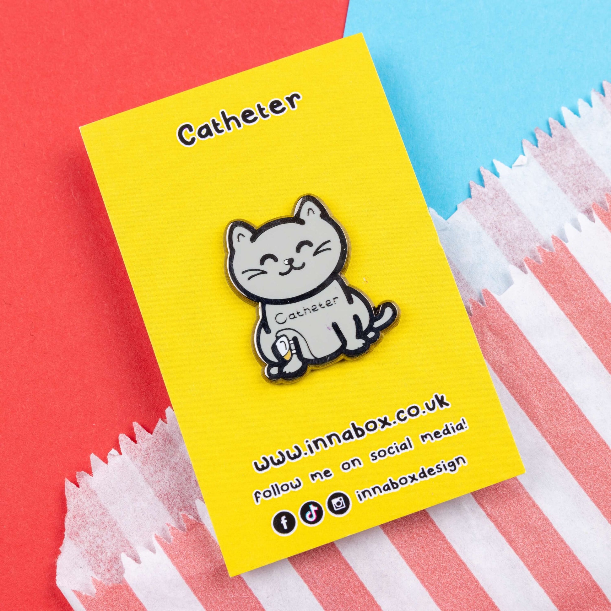 The Catheter Enamel Pin - Catheter on bright yellow backing card laid on a red and blue card background. The pin is a grey smiling cat sat down with a urine drainage Urostomy pouch strapped to its right leg and text across its chest reading 'catheter'. The pin is designed to raise awareness for bladder problems such as UTIs and other chronic illnesses.