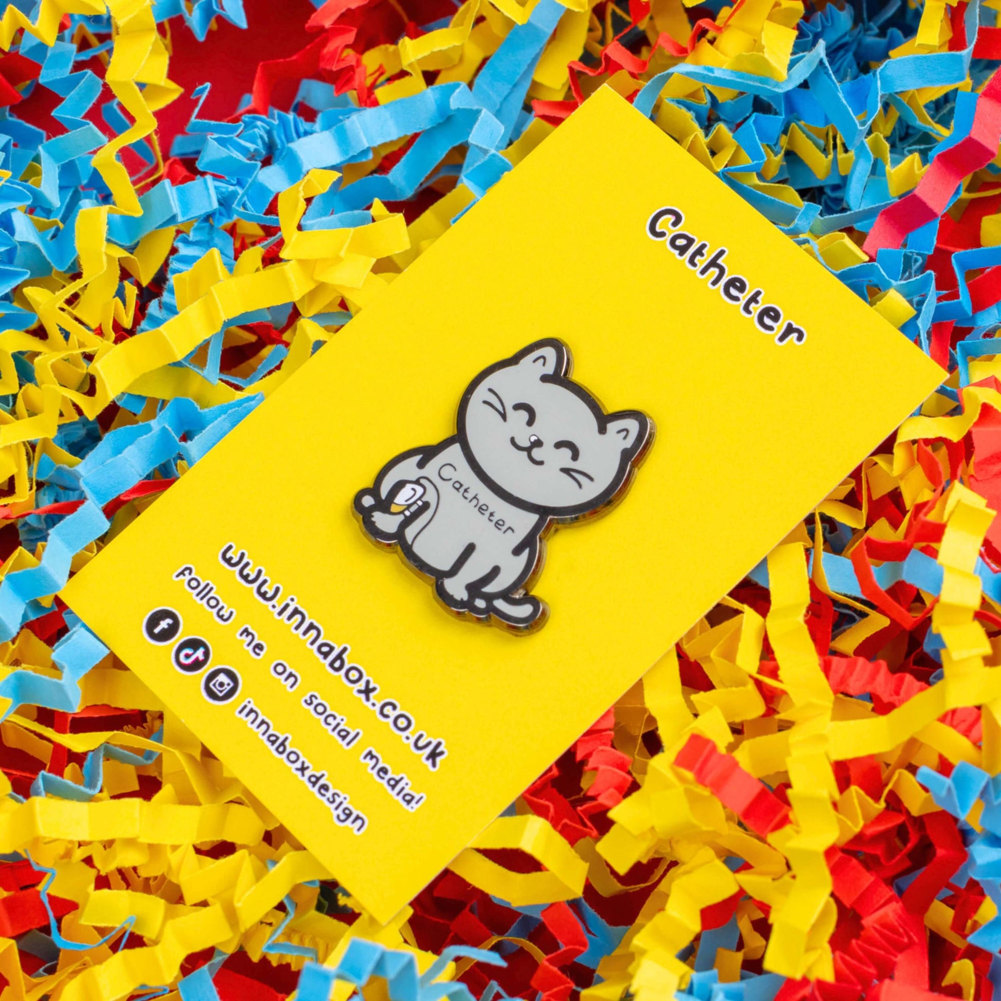 The Catheter Enamel Pin - Catheter on bright yellow backing card laid on a red, yellow and blue card confetti background. The pin is a grey smiling cat sat down with a urine drainage Urostomy pouch strapped to its right leg and text across its chest reading 'catheter'. The pin is designed to raise awareness for bladder problems such as UTIs and other chronic illnesses.