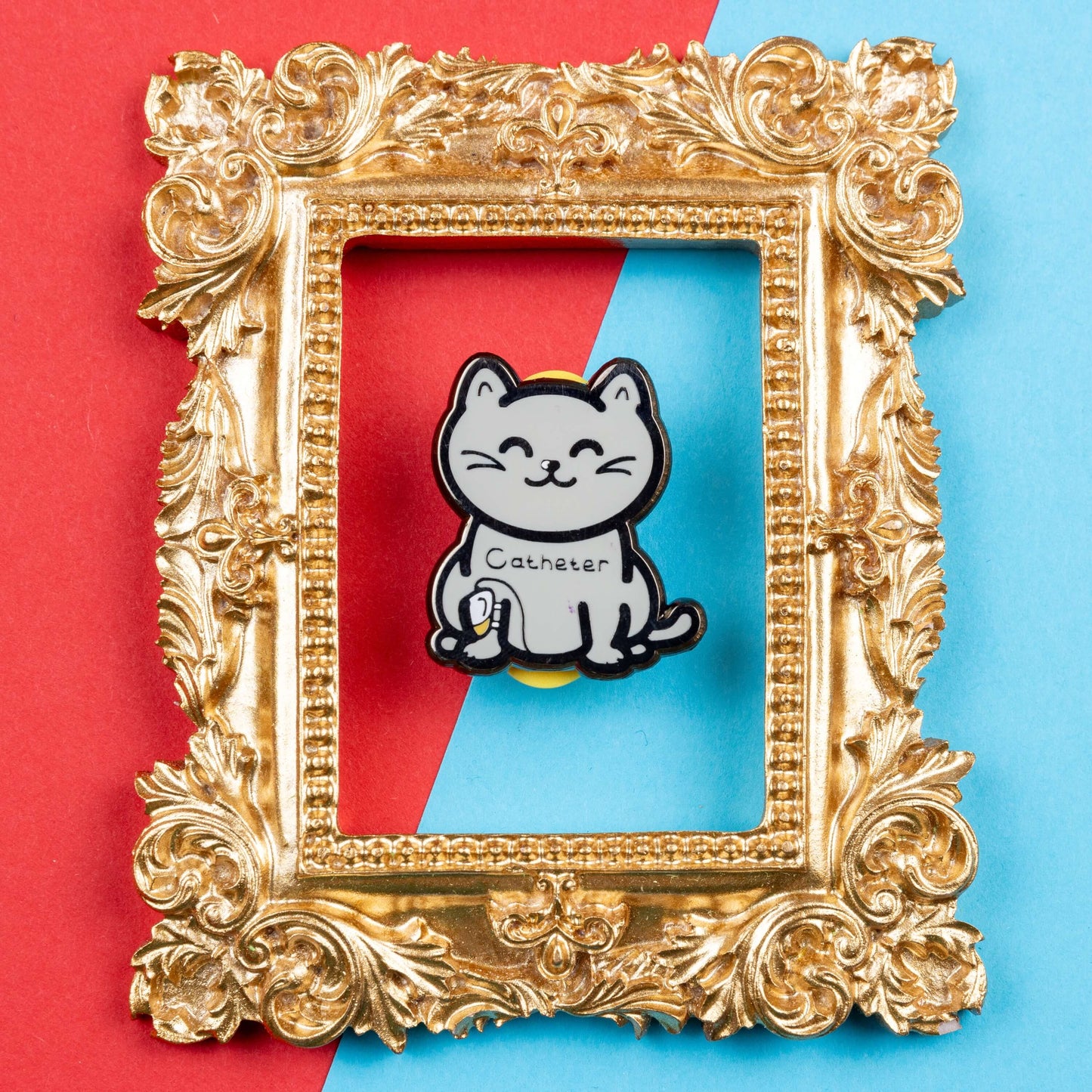 The Catheter Enamel Pin - Catheter on a red and blue card background inside a gold ornate frame. The pin is a grey smiling cat sat down with a urine drainage Urostomy pouch strapped to its right leg and text across its chest reading 'catheter'. The pin is designed to raise awareness for bladder problems such as UTIs and other chronic illnesses.