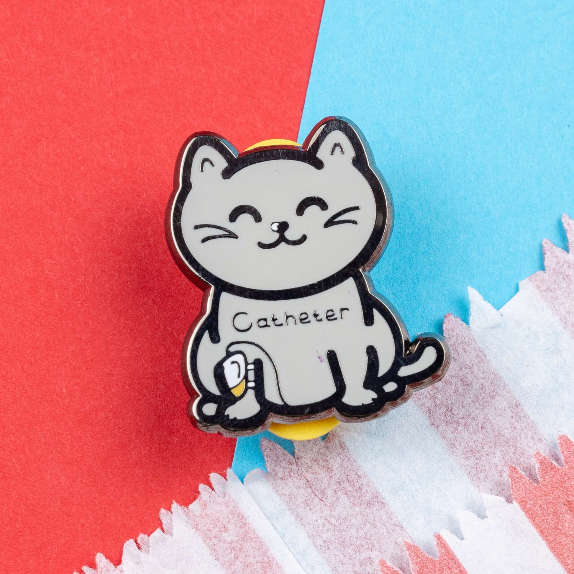 The Catheter Enamel Pin - Catheter on a red and blue card background. The pin is a grey smiling cat sat down with a urine drainage Urostomy pouch strapped to its right leg and text across its chest reading 'catheter'. The pin is designed to raise awareness for bladder problems such as UTIs and other chronic illnesses.