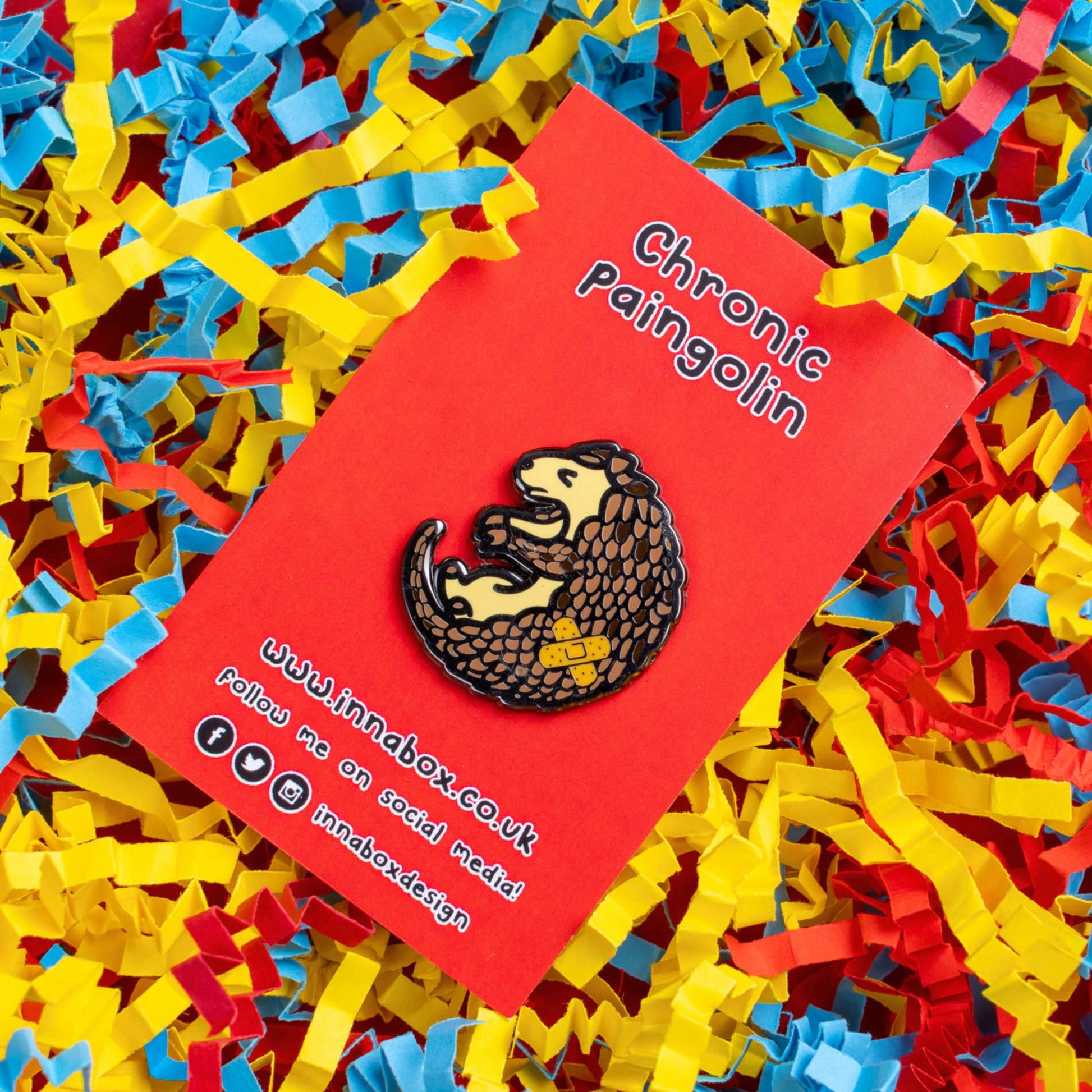 The Chronic Paingolin Enamel Pin - Chronic Pain on red backing card laid on blue, yellow and red card confetti. The pangolin shaped pin has a frustrated expression whilst curled up with a double cross bandaid plaster on its side. The design was created to raise awareness for chronic pain.