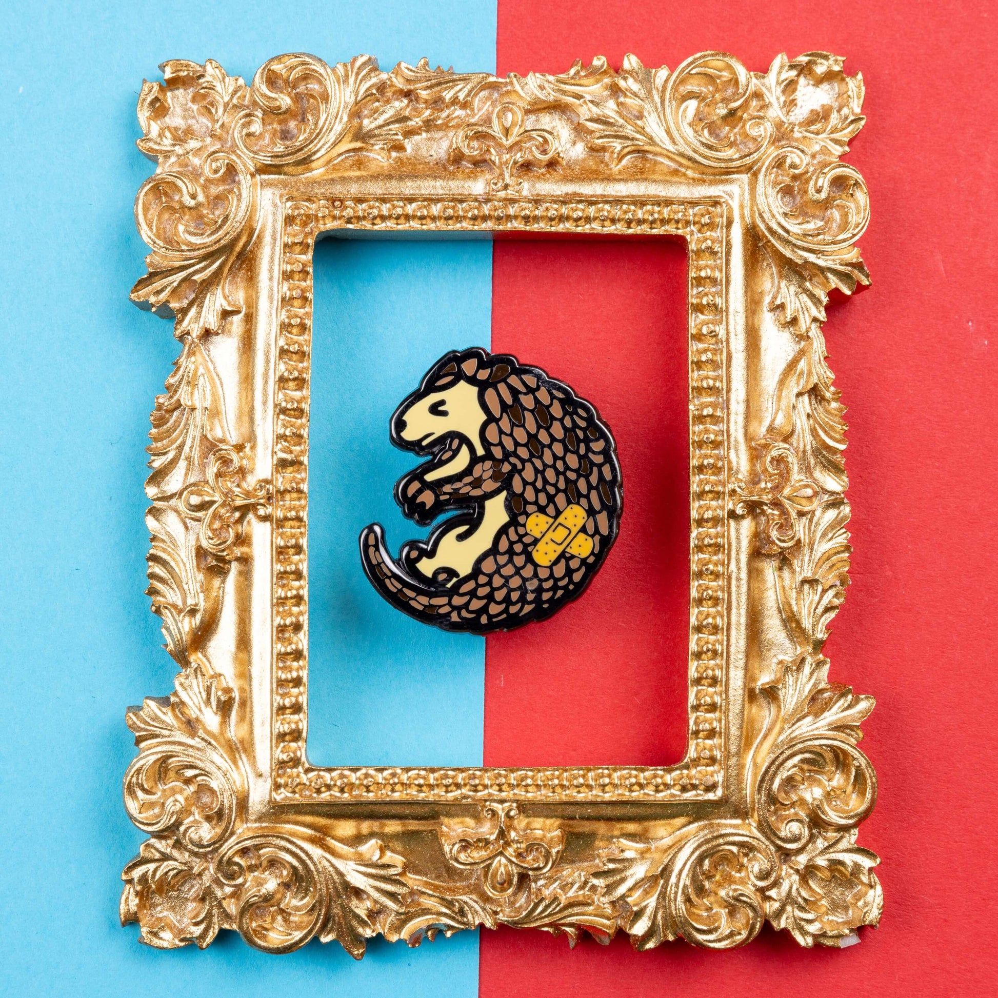 The Chronic Paingolin Enamel Pin - Chronic Pain on blue and red card inside a gold ornate frame. The pangolin shaped pin has a frustrated expression whilst curled up with a double cross bandaid plaster on its side. The design was created to raise awareness for chronic pain.