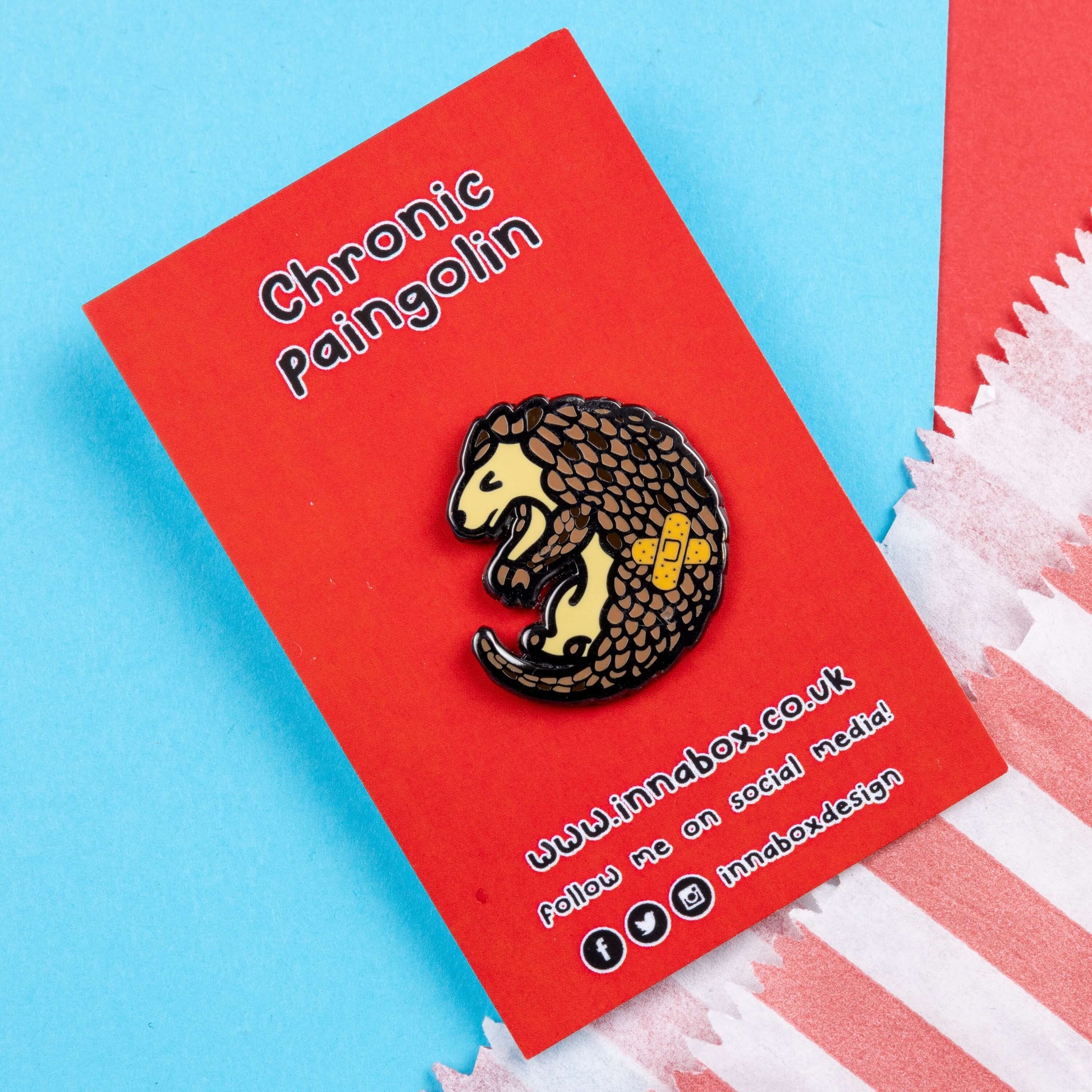 The Chronic Paingolin Enamel Pin - Chronic Pain on red backing card laid on blue and red card. The pangolin shaped pin has a frustrated expression whilst curled up with a double cross bandaid plaster on its side. The design was created to raise awareness for chronic pain.