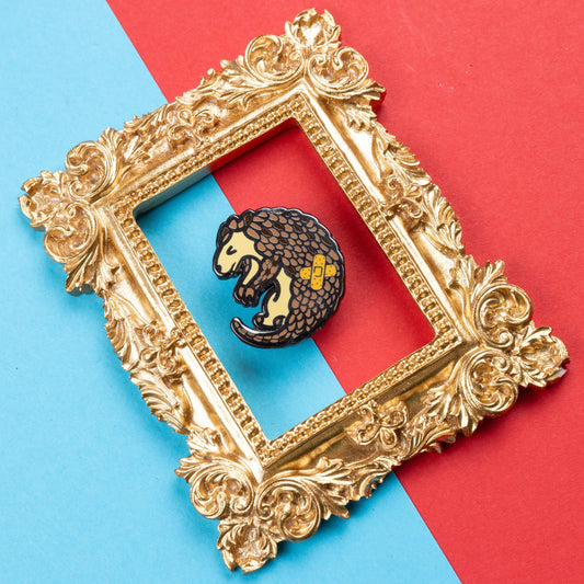 The Chronic Paingolin Enamel Pin - Chronic Pain on blue and red card inside a gold ornate frame. The pangolin shaped pin has a frustrated expression whilst curled up with a double cross bandaid plaster on its side. The design was created to raise awareness for chronic pain.