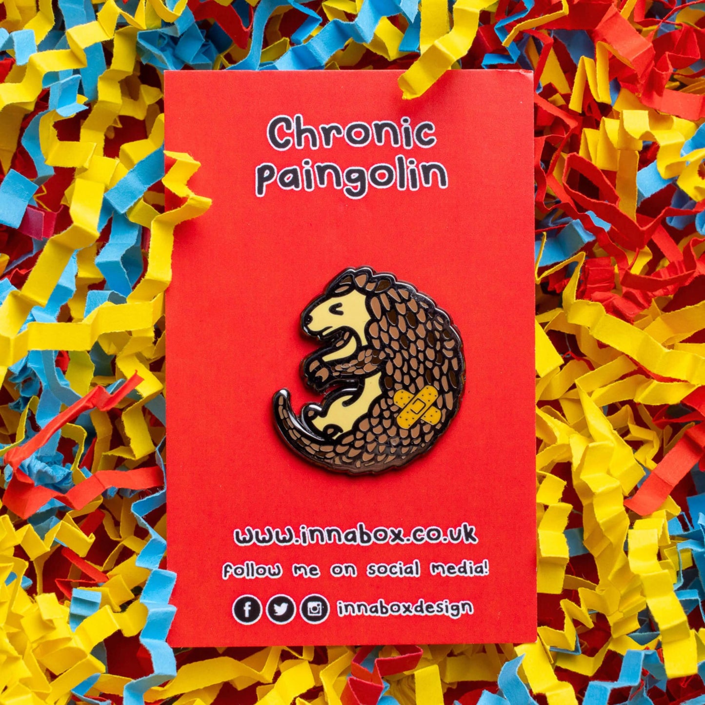 The Chronic Paingolin Enamel Pin - Chronic Pain on red backing card laid on blue, yellow and red card confetti. The pangolin shaped pin has a frustrated expression whilst curled up with a double cross bandaid plaster on its side. The design was created to raise awareness for chronic pain.