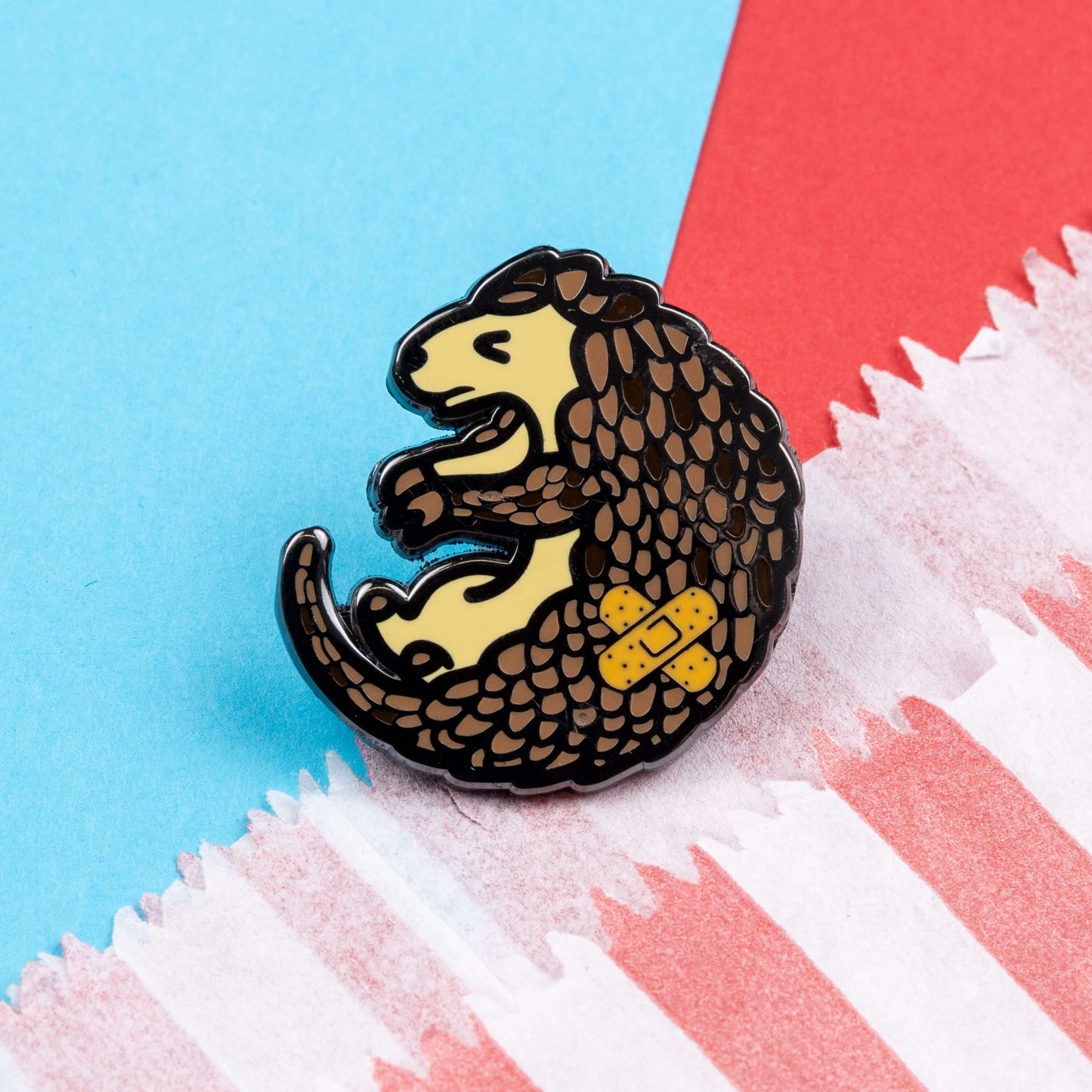 The Chronic Paingolin Enamel Pin - Chronic Pain on blue and red card. The pangolin shaped pin has a frustrated expression whilst curled up with a double cross bandaid plaster on its side. The design was created to raise awareness for chronic pain.