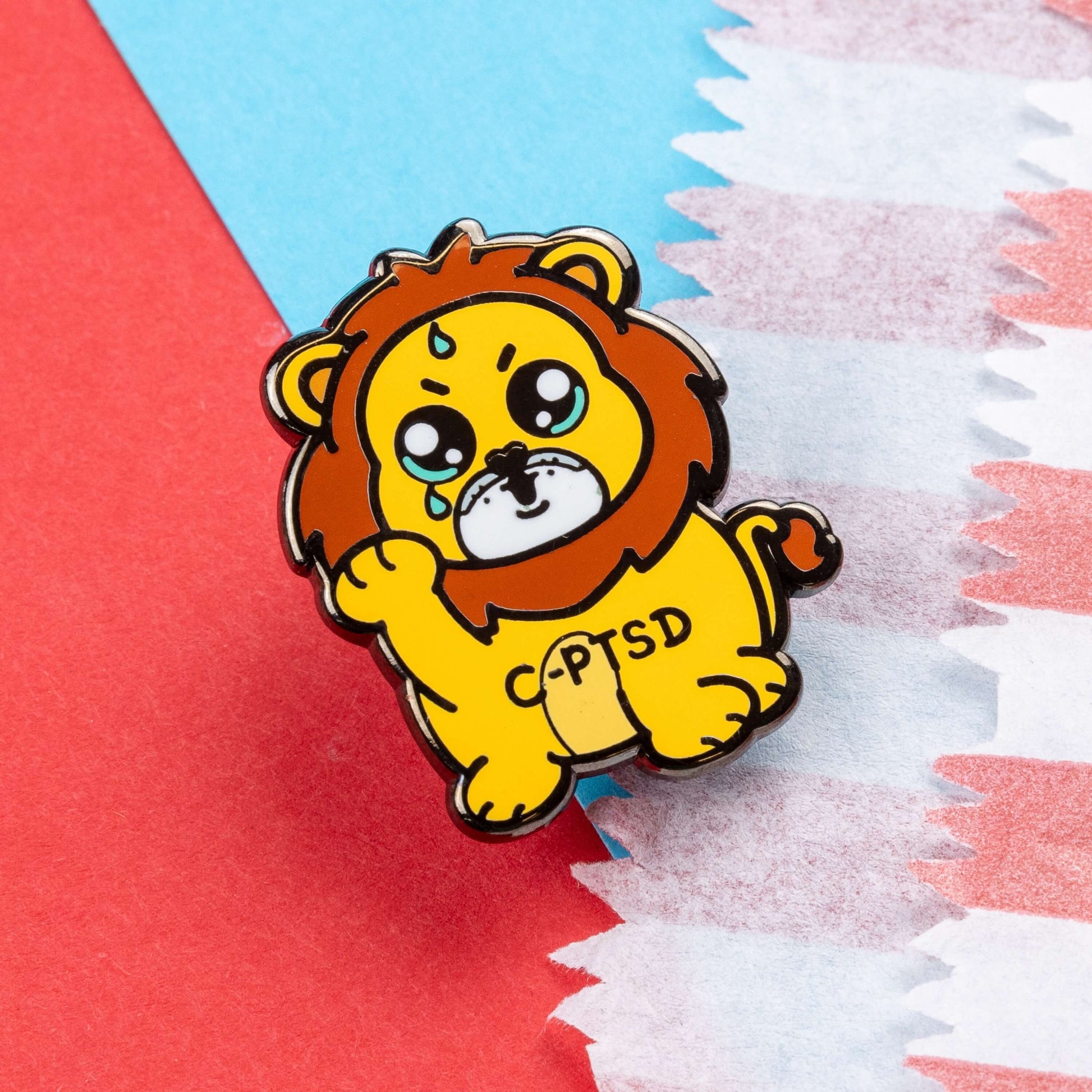 The Complex Post Troarmatic Stress Disorder Lion Enamel Pin - C-PTSD - Complex Post Traumatic Stress Disorder on a red and blue card background. A yellow crying smiling and sweating male lion with its paw raised in the middle with 'C-PTSD' across its chest. The enamel pin design is raising awareness for complex post traumatic stress disorder.