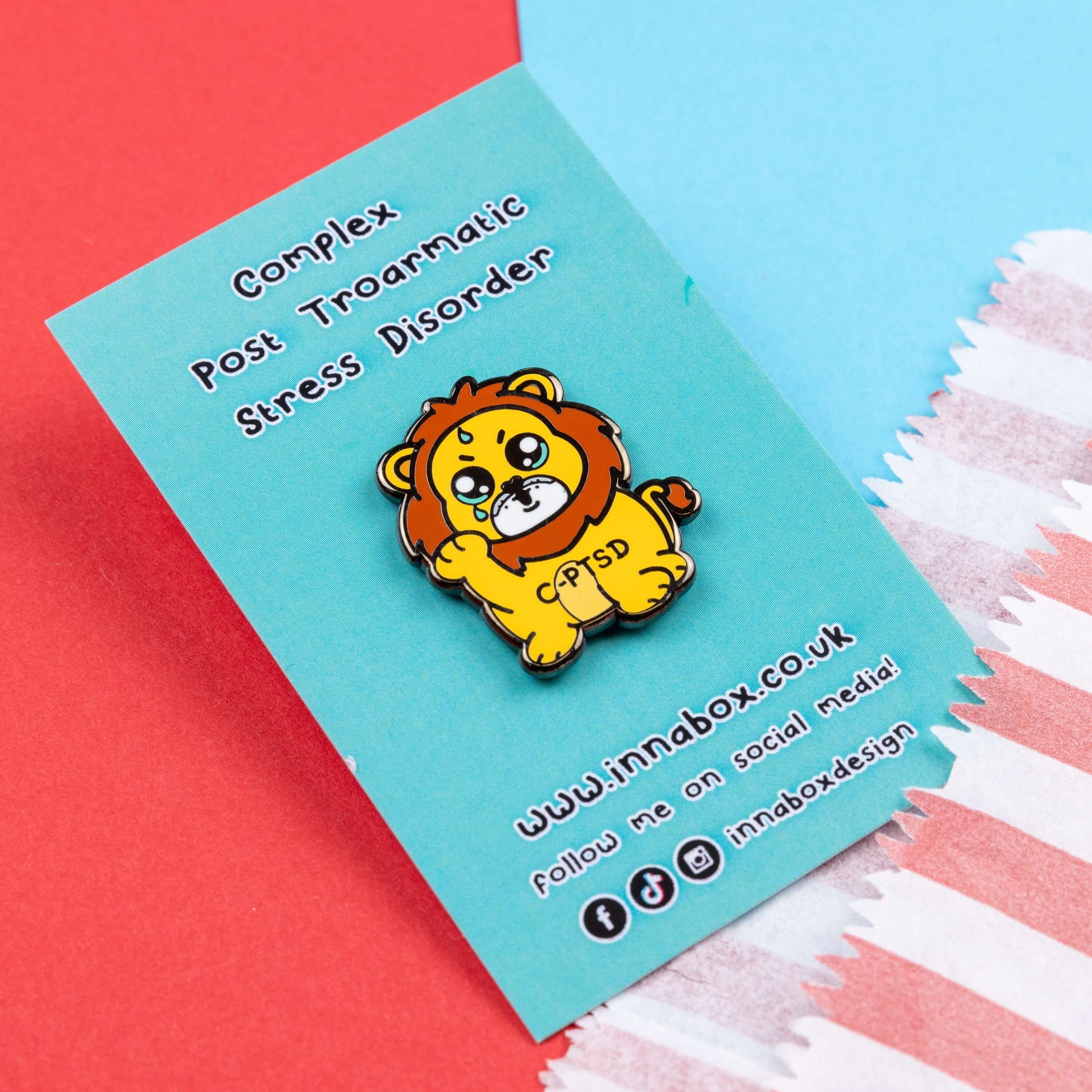 The Complex Post Troarmatic Stress Disorder Lion Enamel Pin - C-PTSD - Complex Post Traumatic Stress Disorder on blue backing card with black text on blue and red card. A yellow crying smiling and sweating male lion with its paw raised in the middle with 'C-PTSD' across its chest. The enamel pin design is raising awareness for complex post traumatic stress disorder.