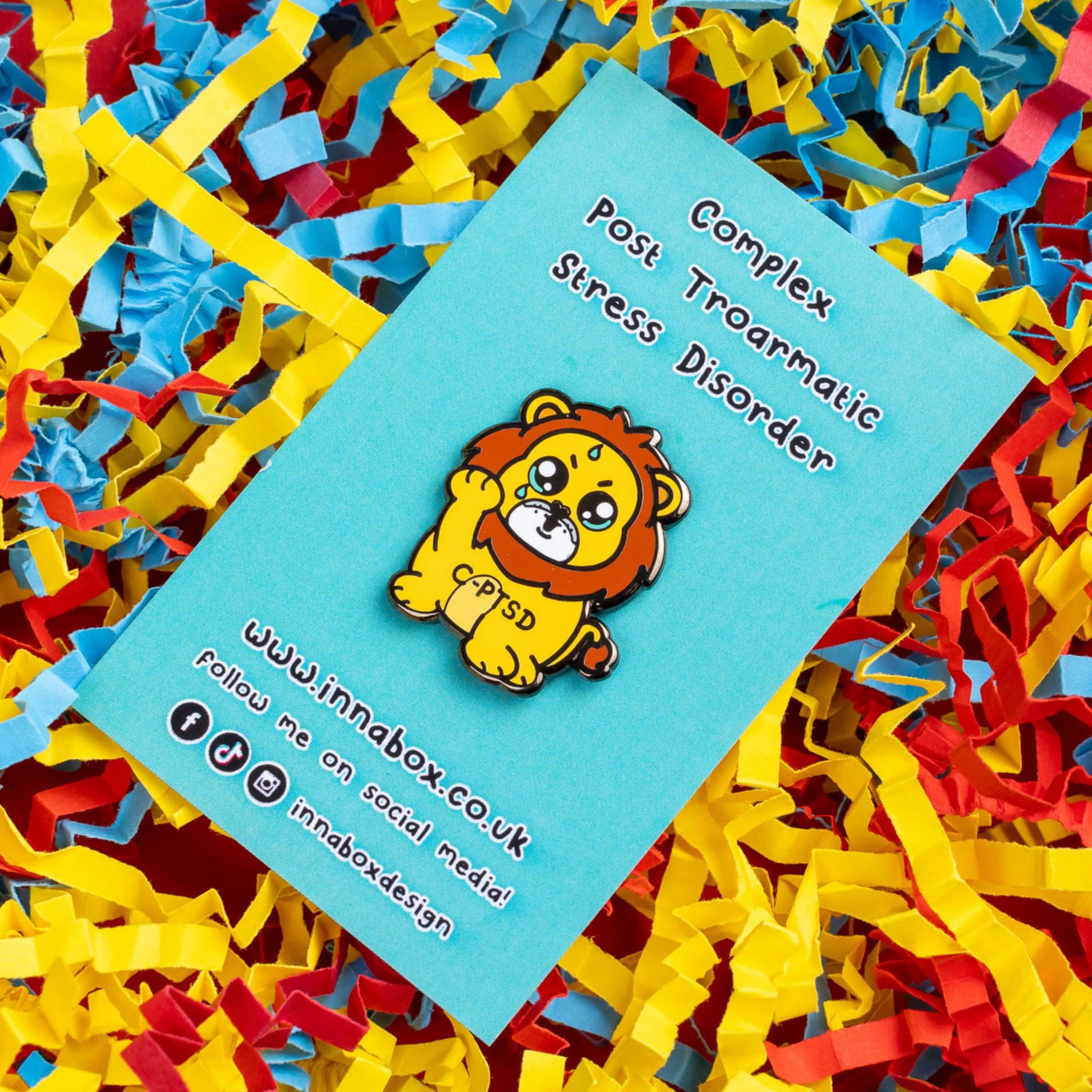 The Complex Post Troarmatic Stress Disorder Lion Enamel Pin - C-PTSD - Complex Post Traumatic Stress Disorder on blue backing card laid on a red, yellow and blue card confetti background. A yellow crying smiling and sweating male lion with its paw raised in the middle with 'C-PTSD' across its chest. The enamel pin design is raising awareness for complex post traumatic stress disorder.